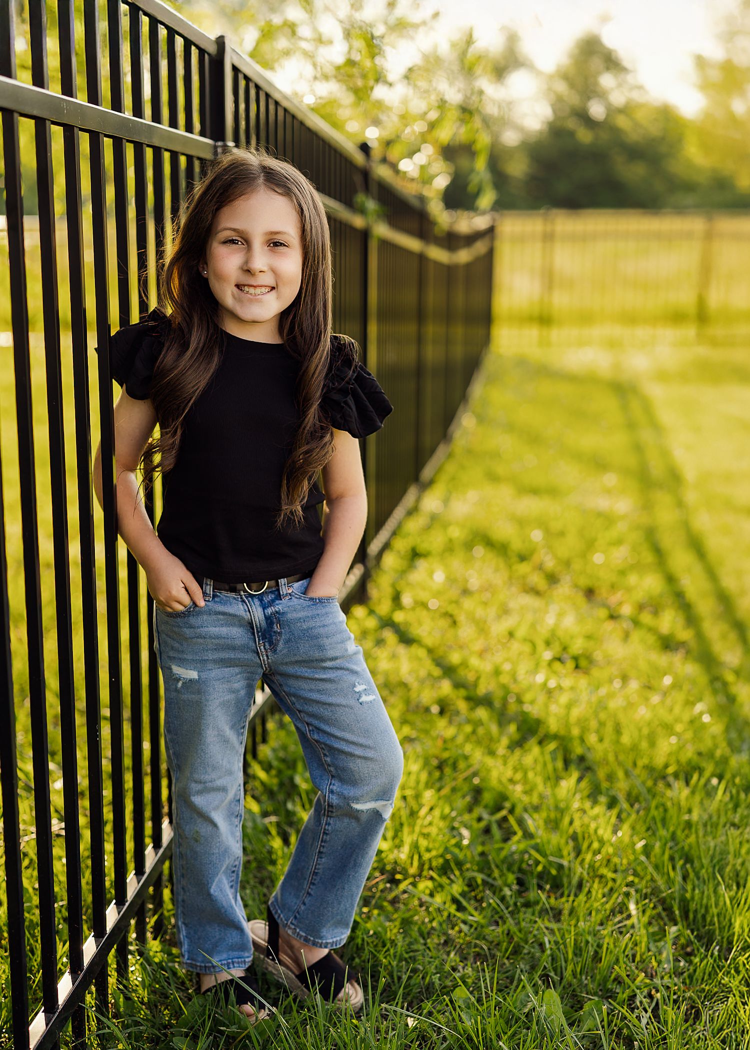 Child wearing casual outfit stands near black metal fence in golden evening light.