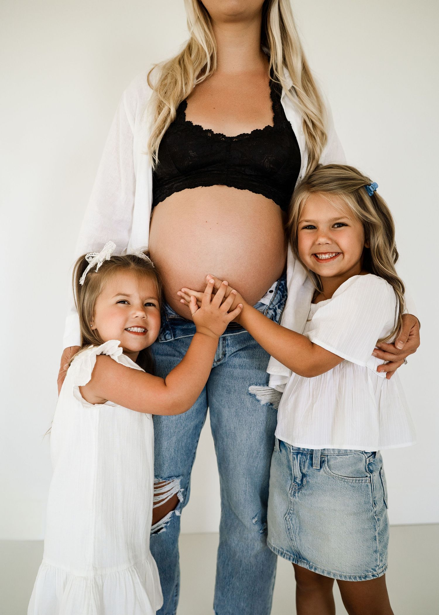 Family maternity photo with children hugging pregnant belly in casual denim and white outfits.