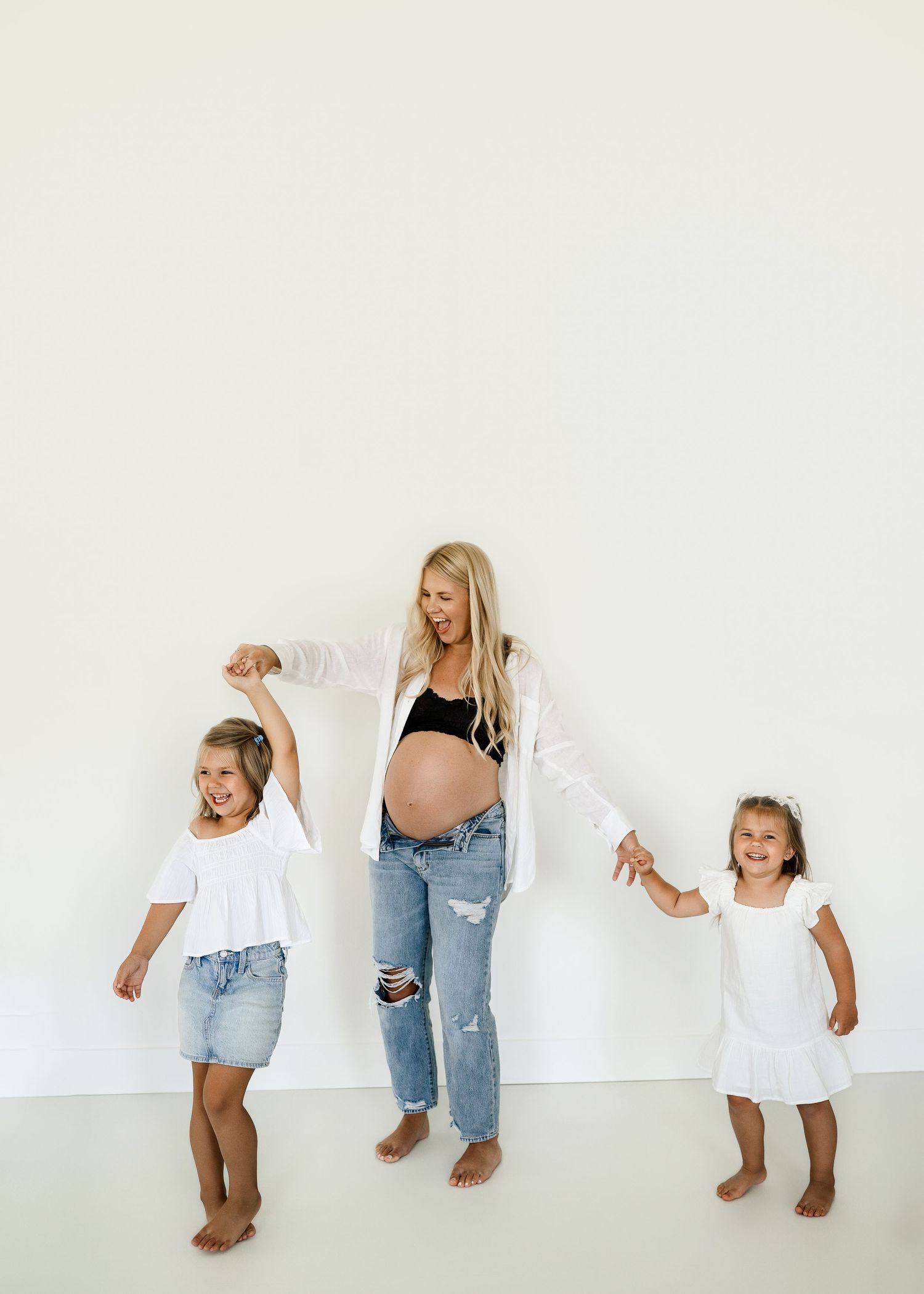 Playful maternity photo of mother and daughters dancing against white wall.