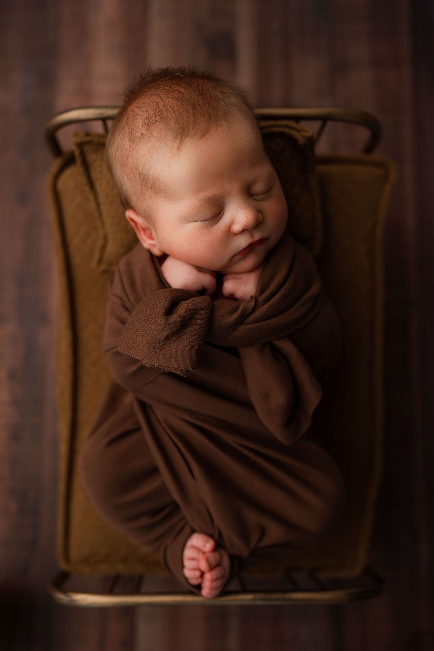 Sleeping newborn baby wrapped in brown blanket resting in vintage chair during professional photo session.