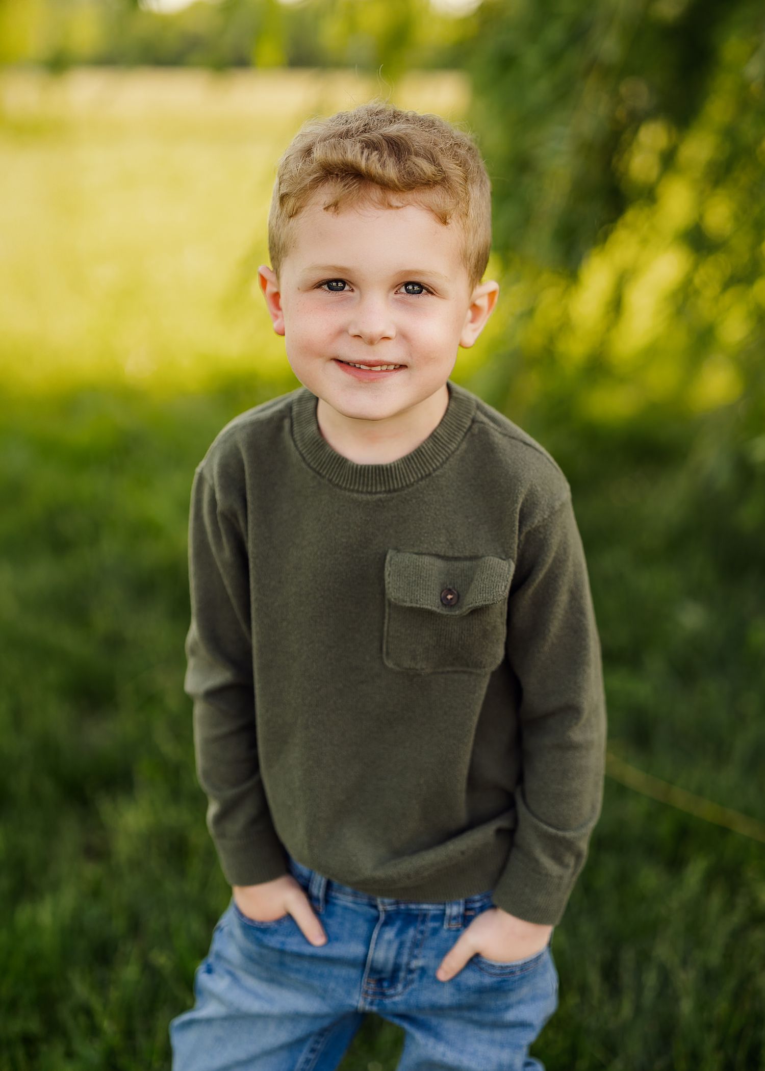 Young child in olive sweater and jeans smiling outdoors in natural setting.