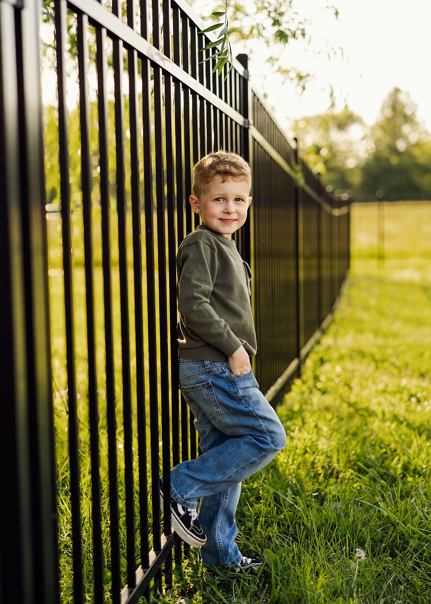 Child in green shirt and jeans poses by metal fence in sunny outdoor setting.