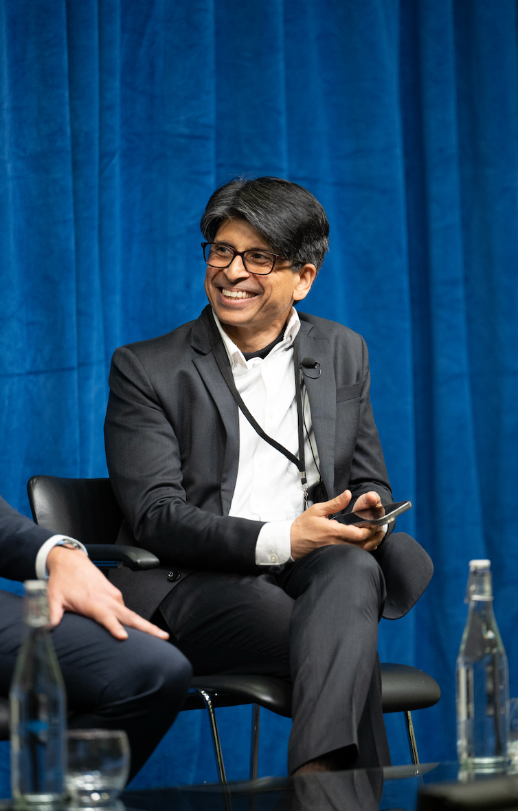 Panel discussion participant in formal attire seated on stage with blue curtain backdrop.
