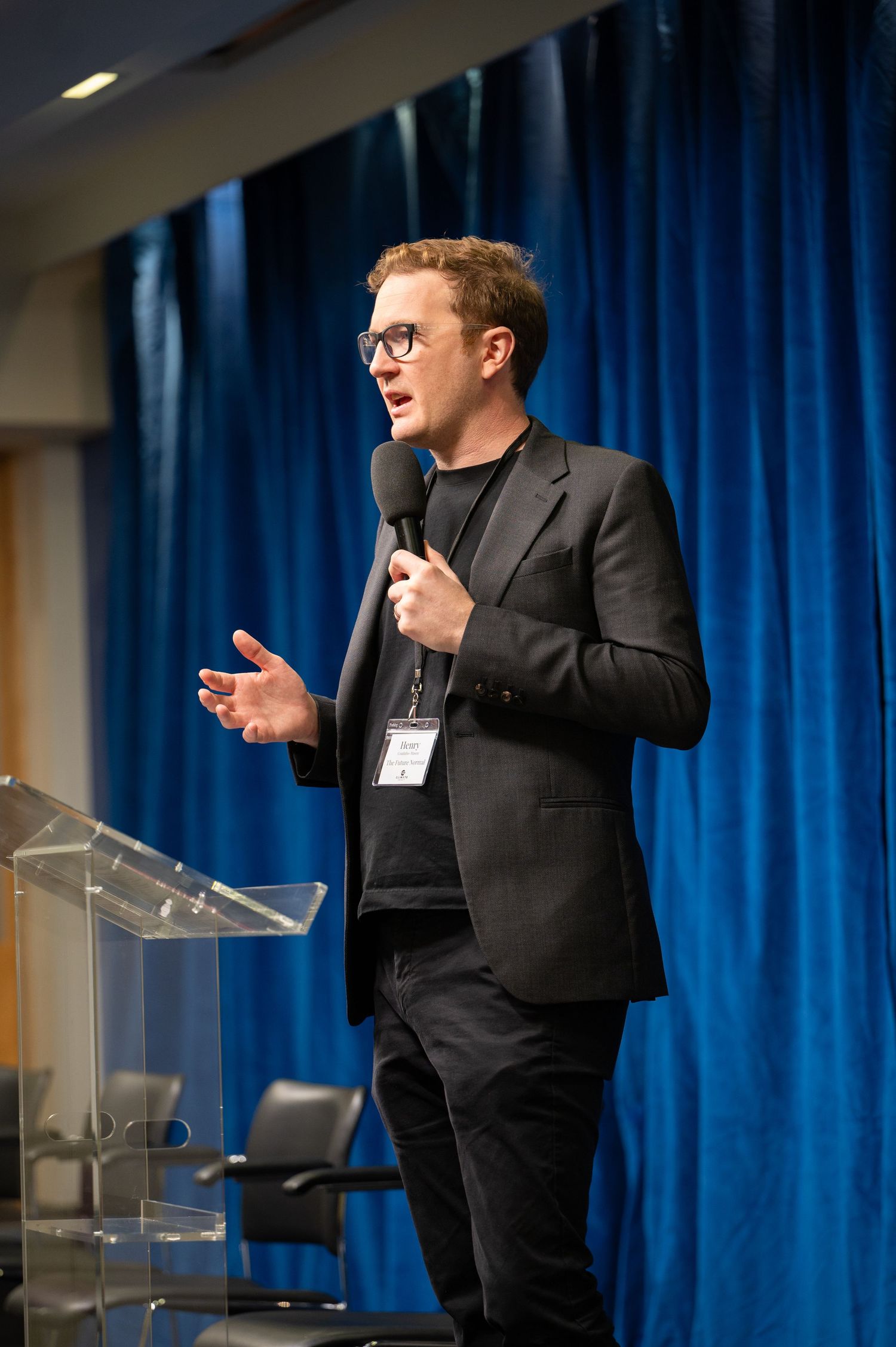 Speaker presenting at a podium with blue curtain backdrop during a professional event.