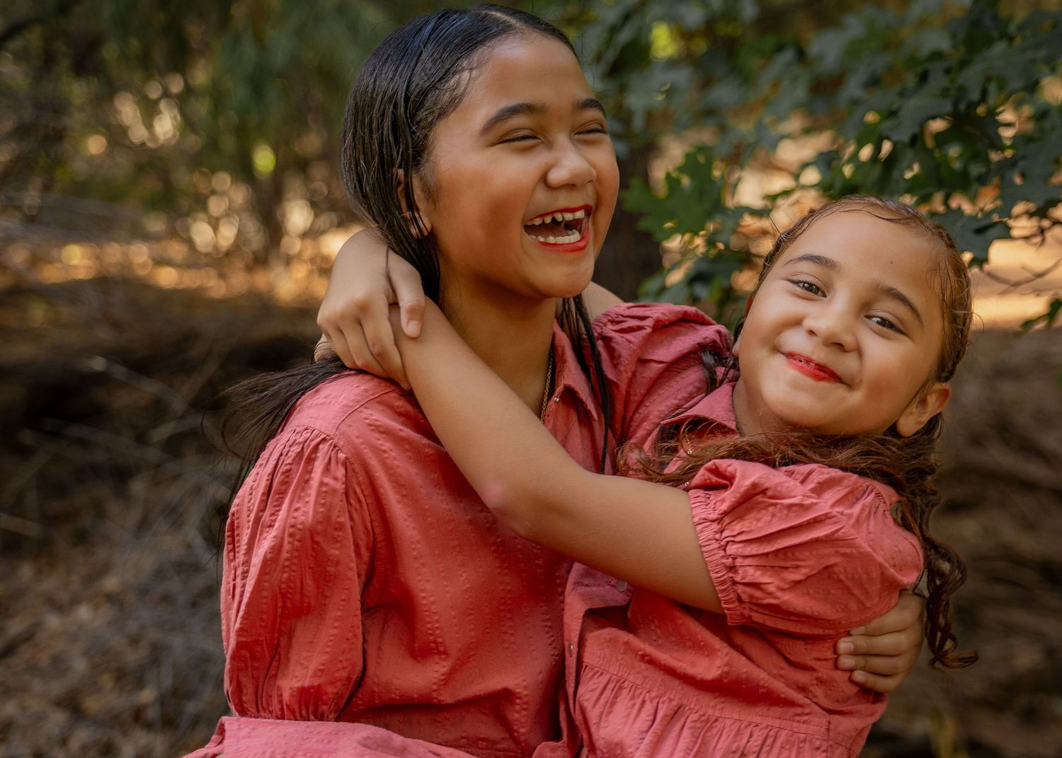 Two young girls in matching red dresses hugging and smiling outdoors among trees.