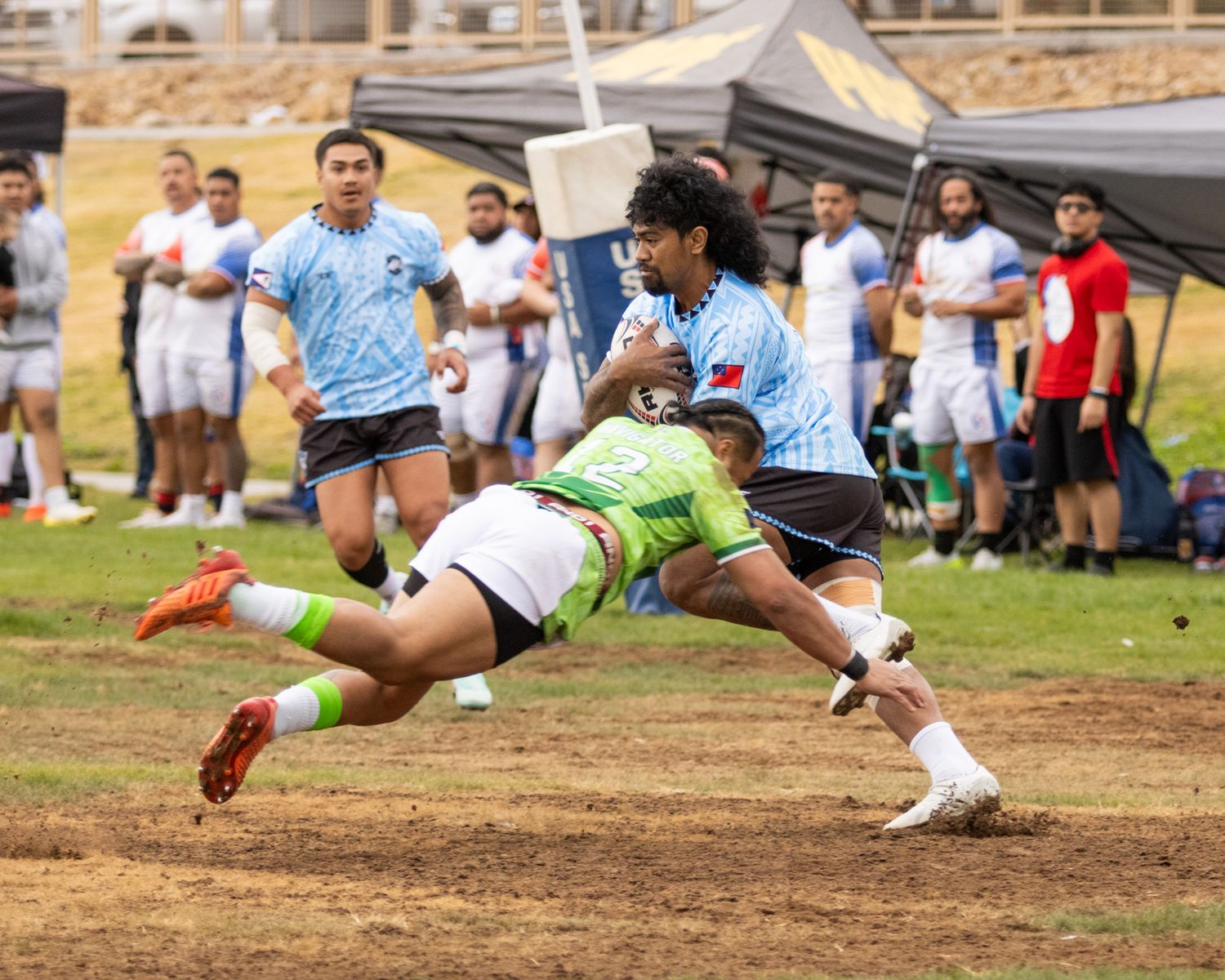 Rugby players compete in an intense tackle during an outdoor match, with teammates and spectators visible in the background.