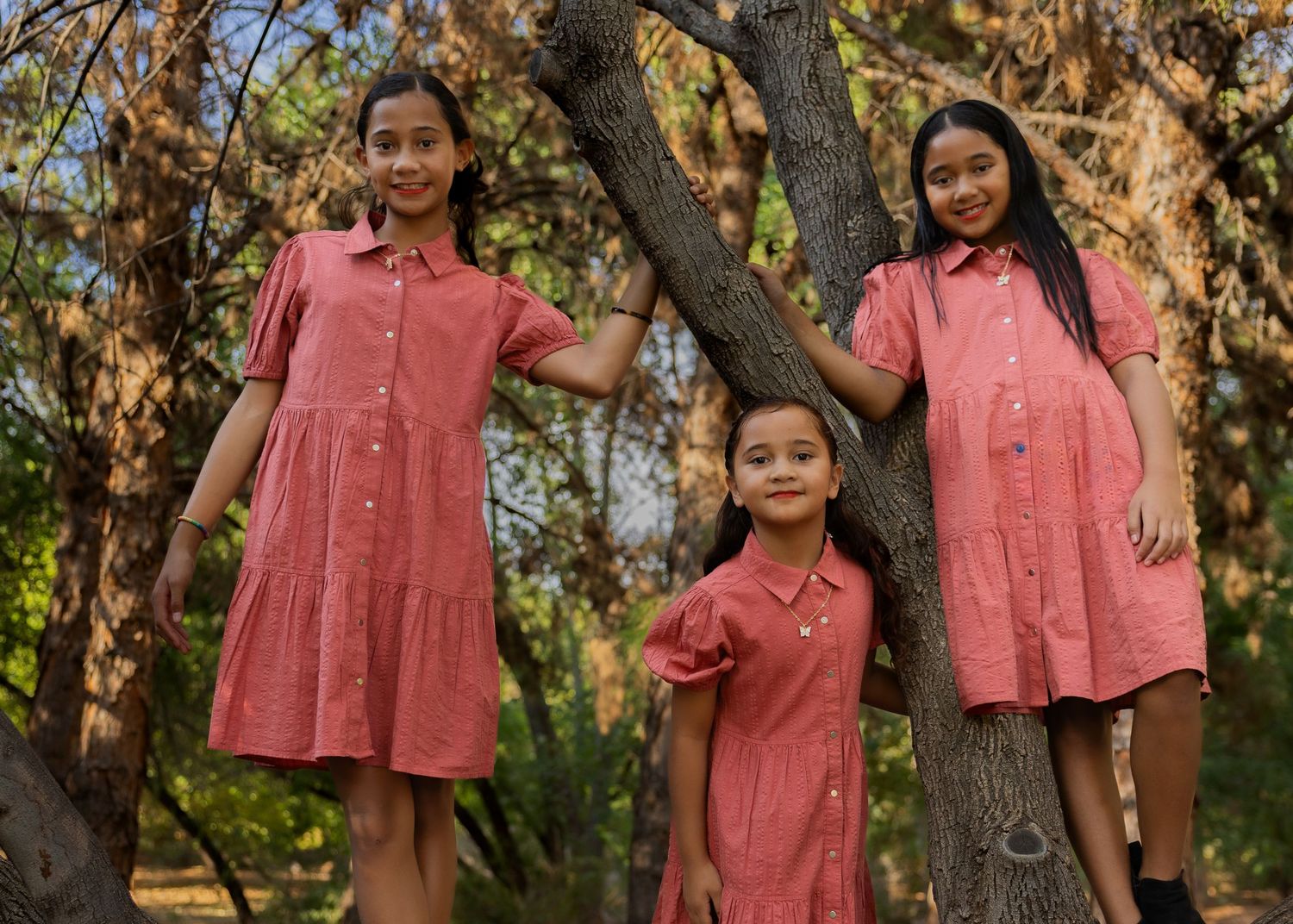 Three girls in matching red button-up dresses pose outdoors among autumn trees.