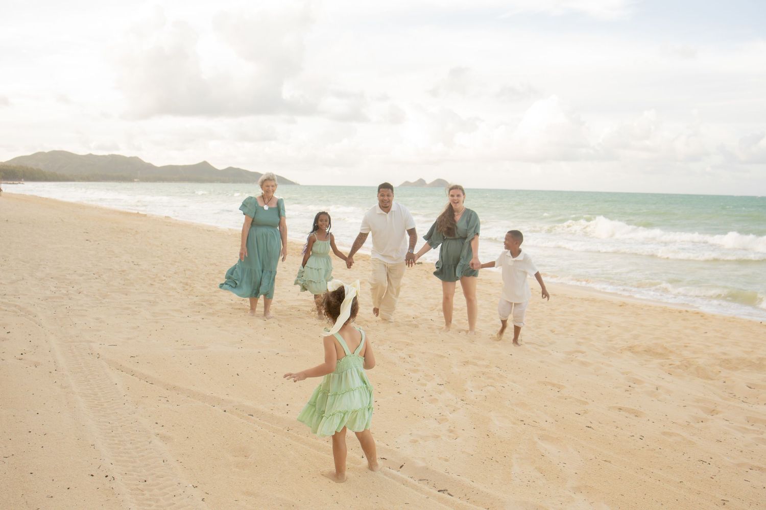 Family in matching teal outfits walking on a sandy beach with turquoise ocean waves and mountains in background.