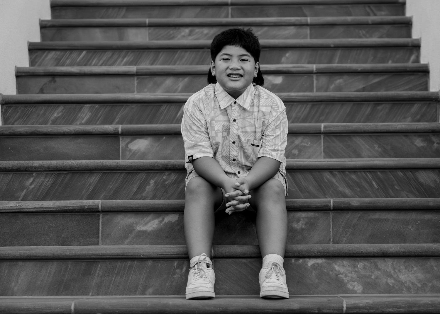 A smiling child sits on stone steps, hands clasped, wearing a patterned shirt and shorts in a black and white photo.