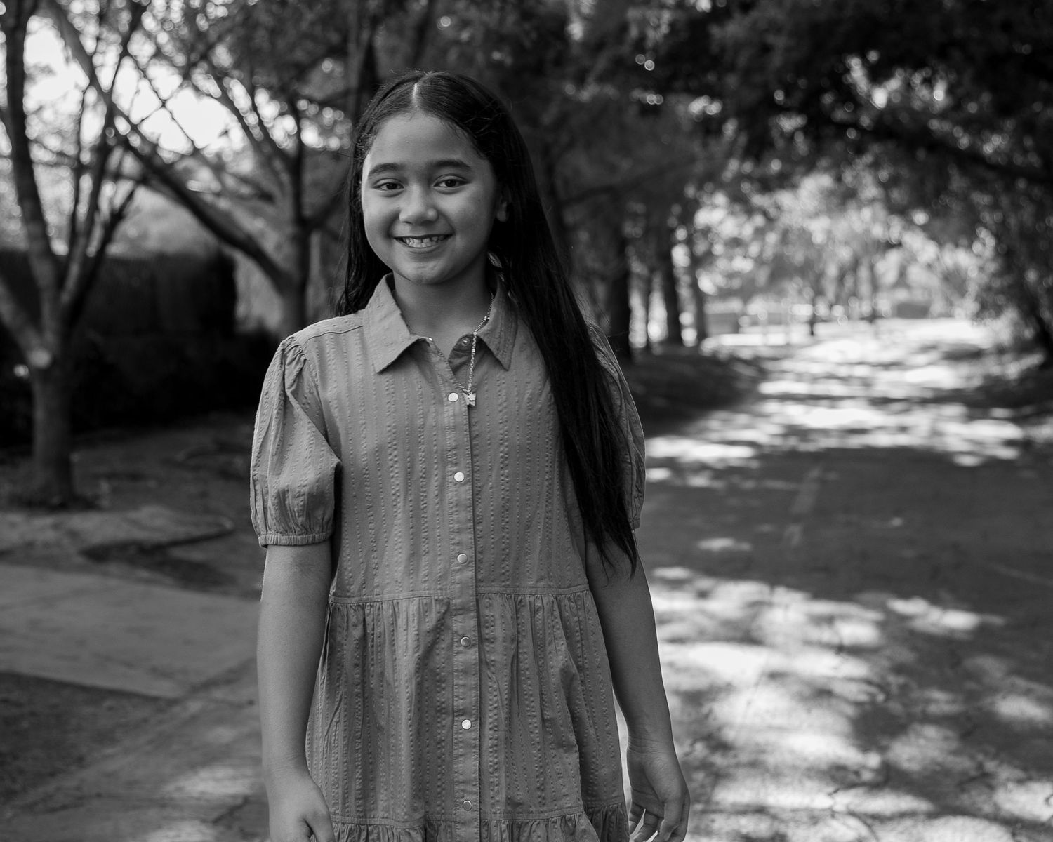 Young girl smiling in a black and white outdoor portrait, wearing a dress, with trees in the background.