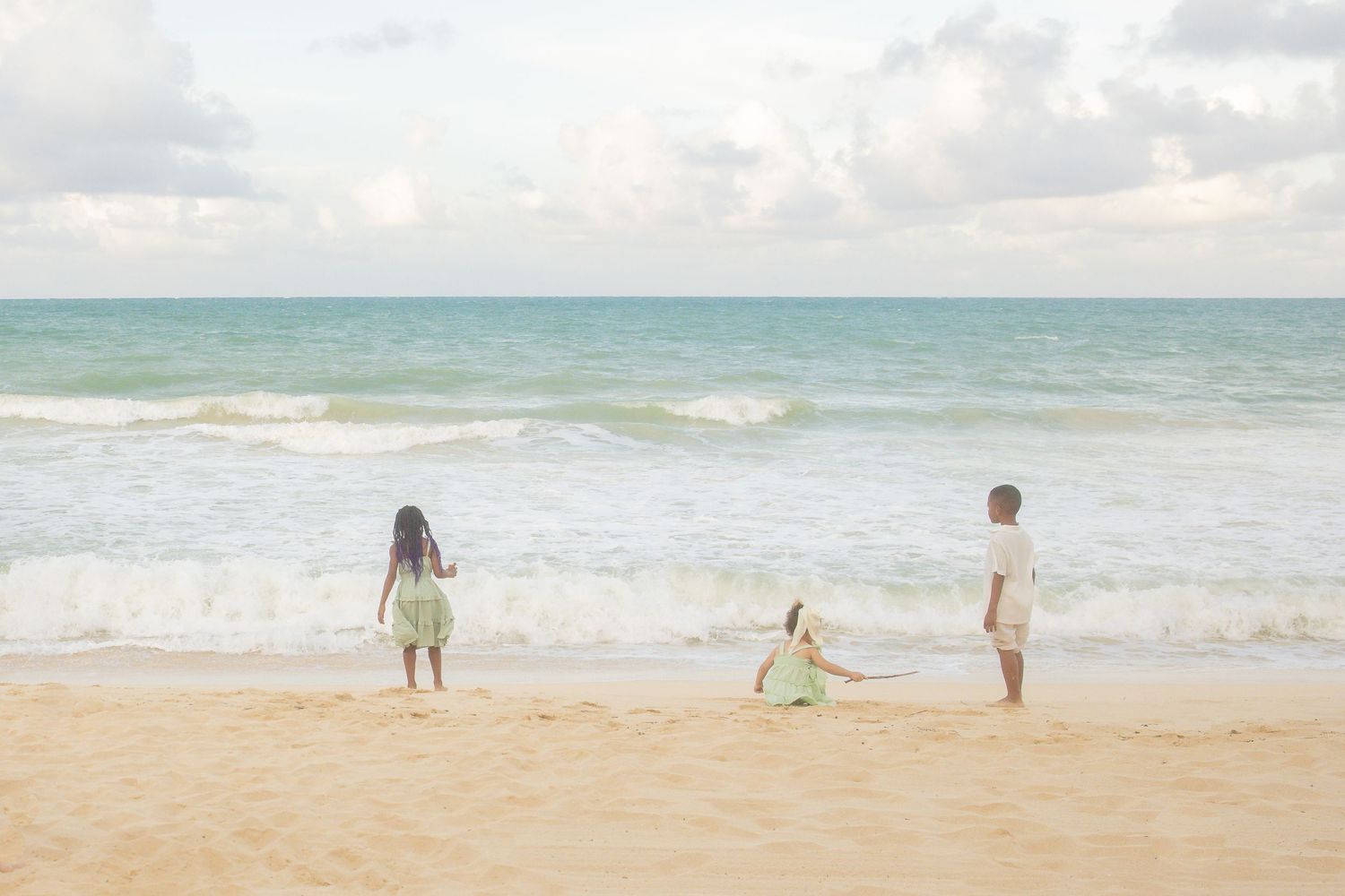 Three children playing on a sandy Hawaiian beach with turquoise ocean waves and cloudy sky in the background.