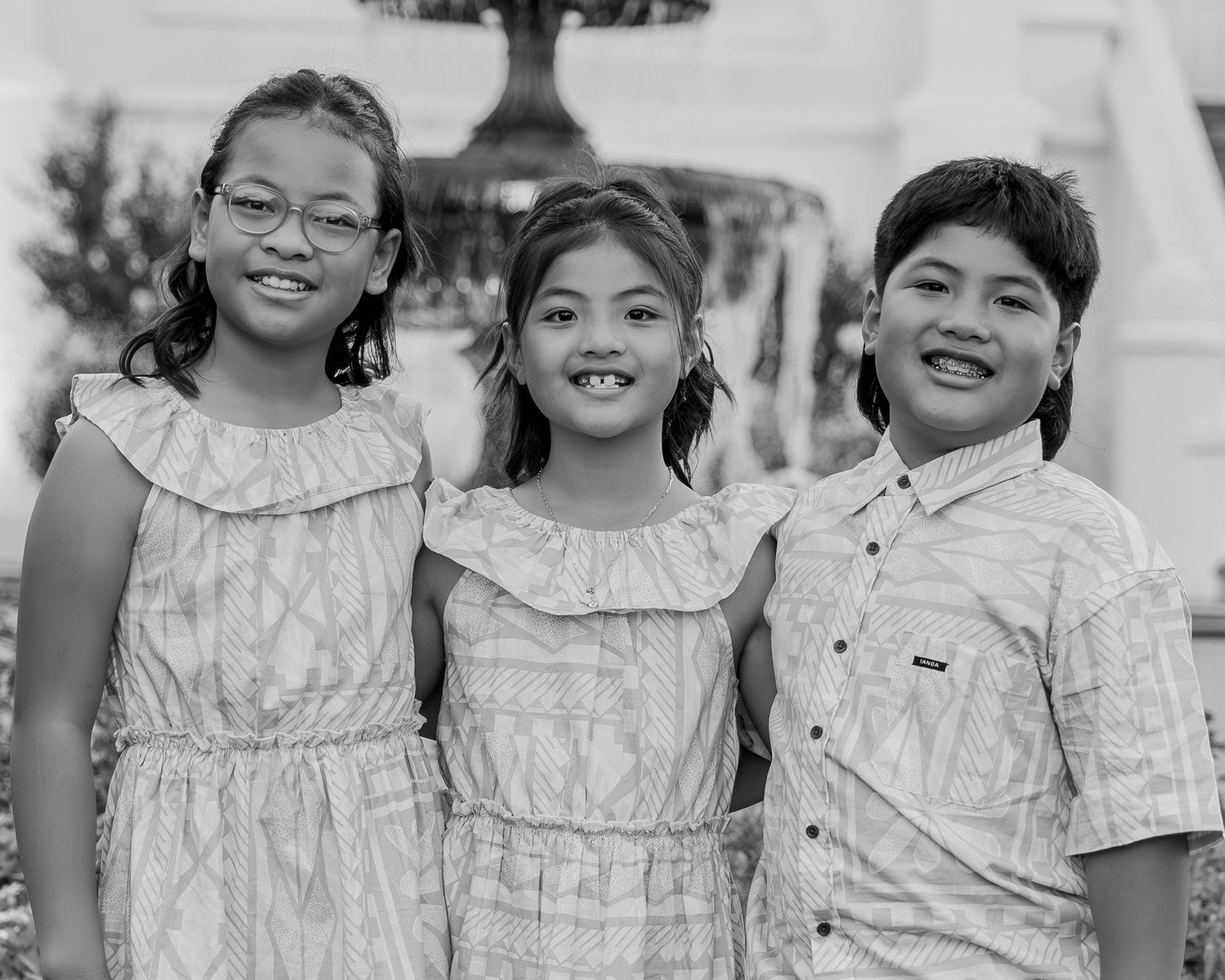 Three smiling children pose together outdoors in black and white photo near a fountain.
