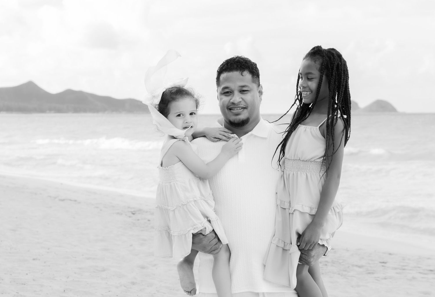 Black and white beach photo of a man holding a toddler beside a woman with braids, ocean and mountains behind.