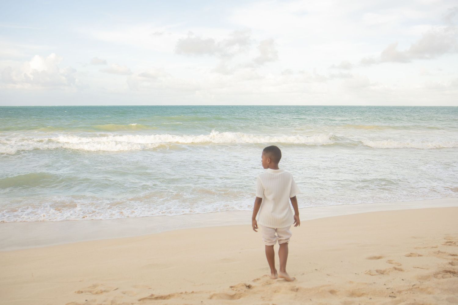 Young child in white outfit standing barefoot on sandy beach gazing at ocean waves under cloudy sky.