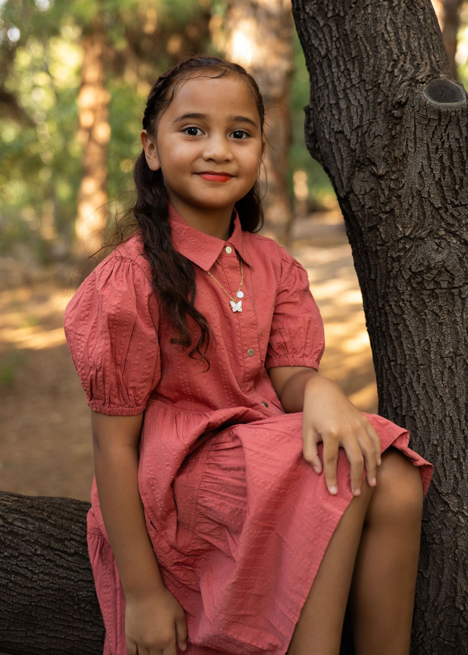 Young girl in pink dress sitting by tree in forest, smiling for portrait photo outdoors.
