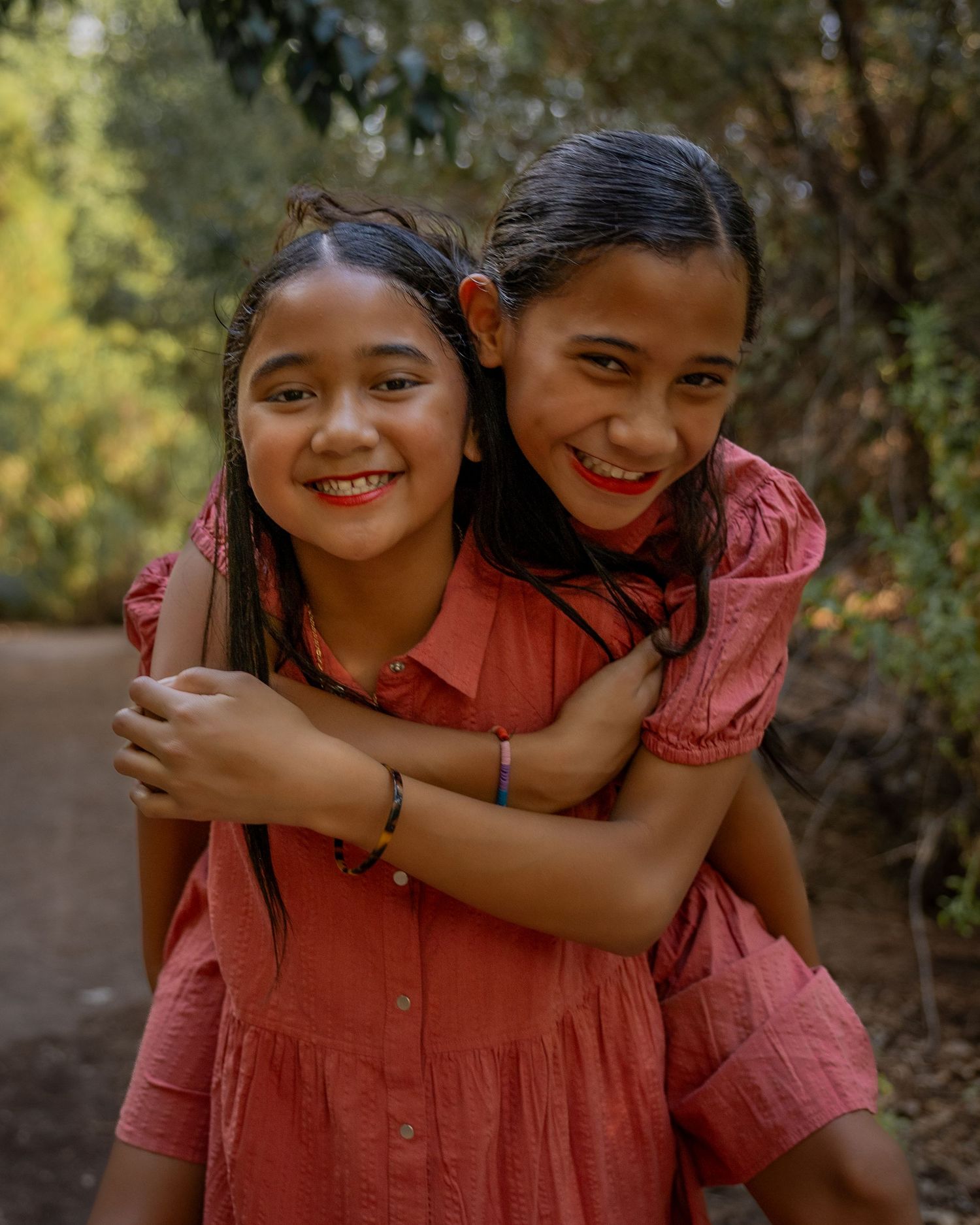 Two smiling girls in matching red dresses share a warm hug outdoors on a tree-lined path.