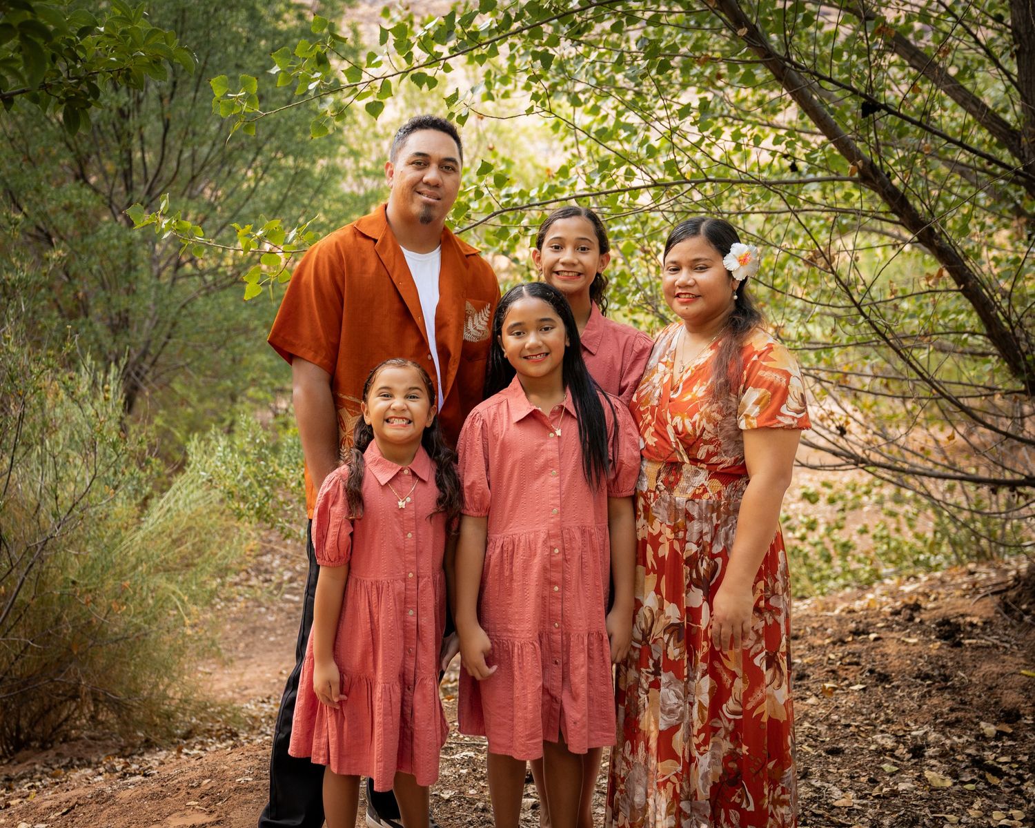 Family of five posing outdoors on a nature trail, wearing coordinating red and coral outfits, surrounded by lush green trees.