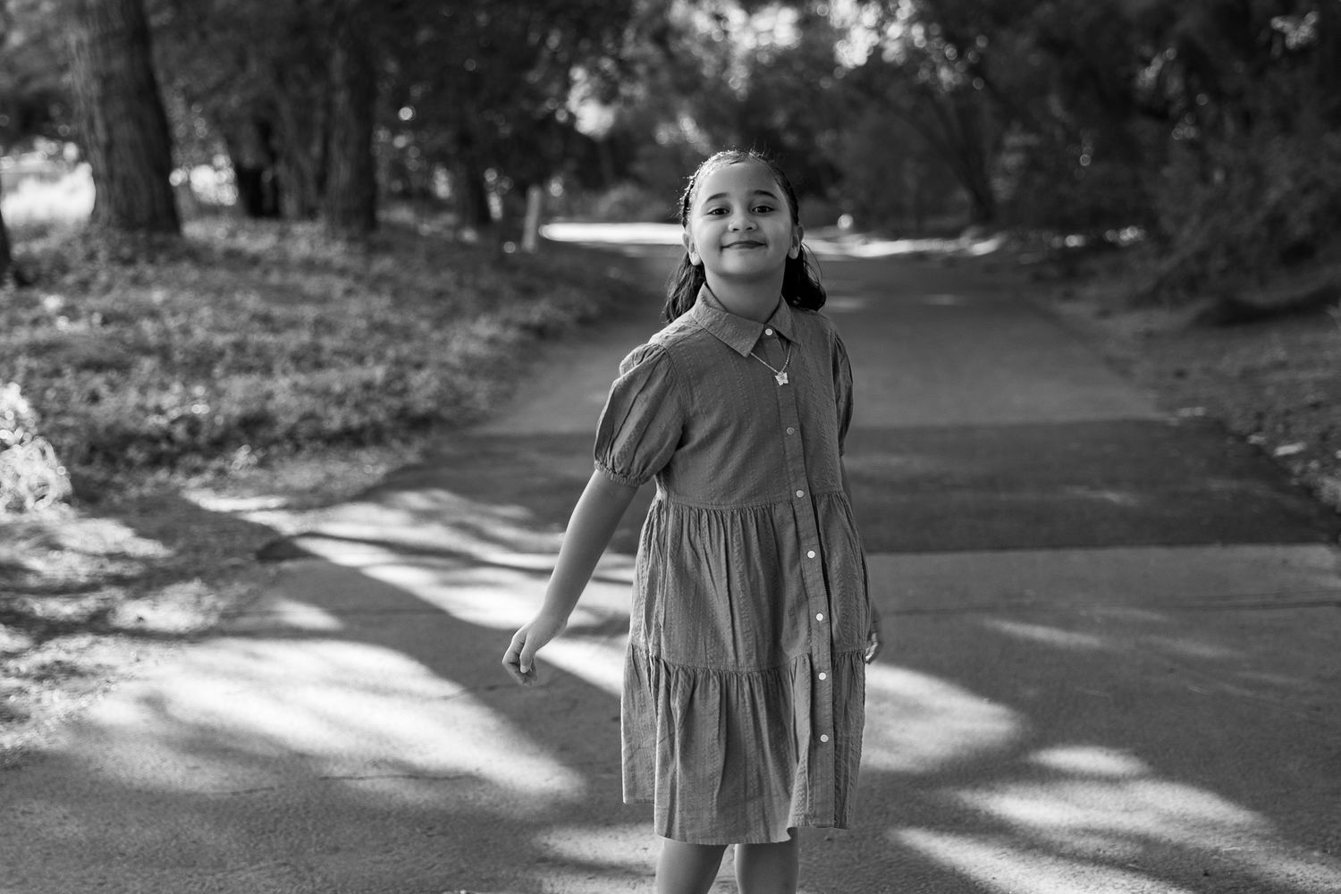 A young girl in a dress smiles joyfully on a tree-lined path in a black and white photo.
