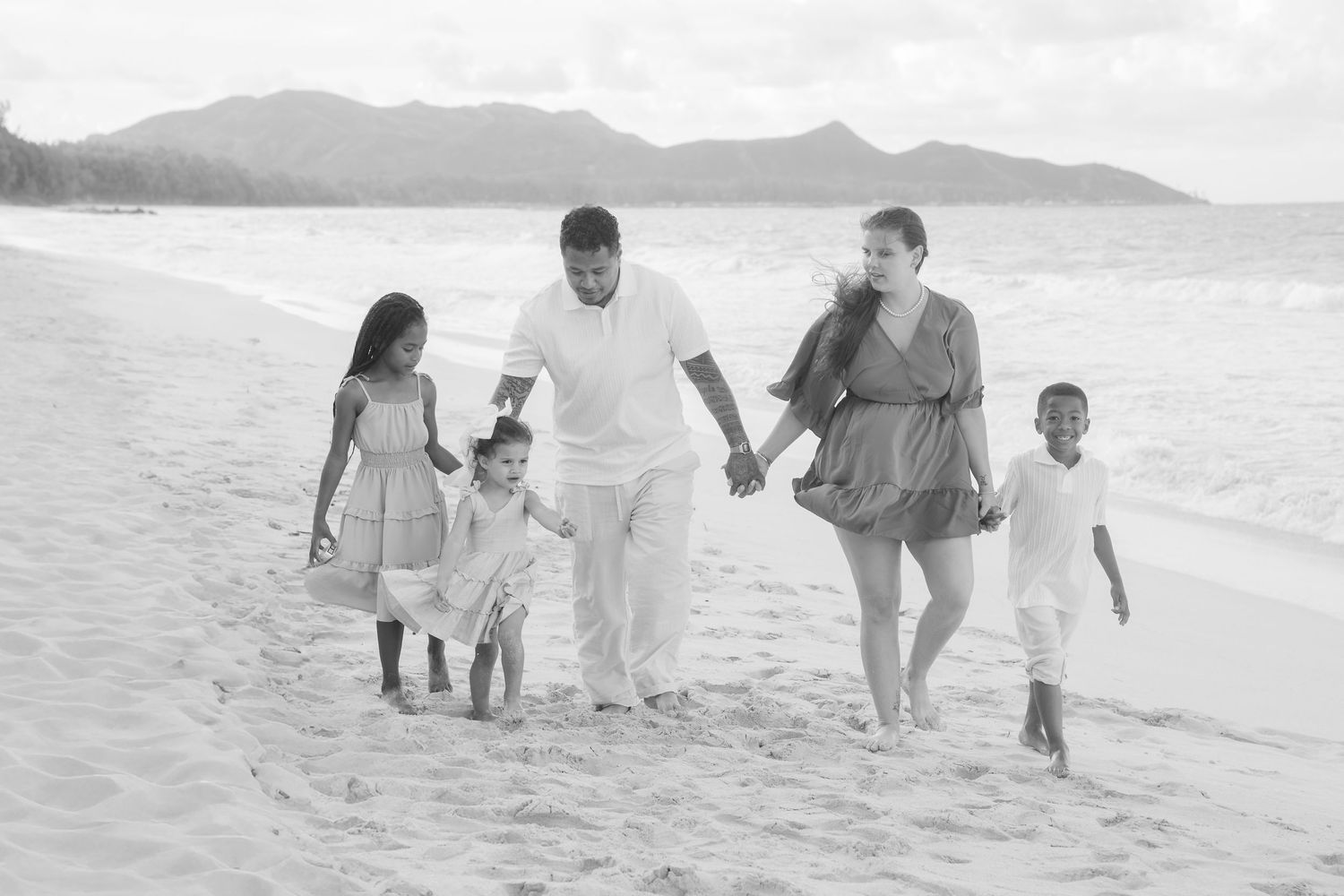 Black and white photo of a happy family of five walking on a sandy beach with mountains in the background.