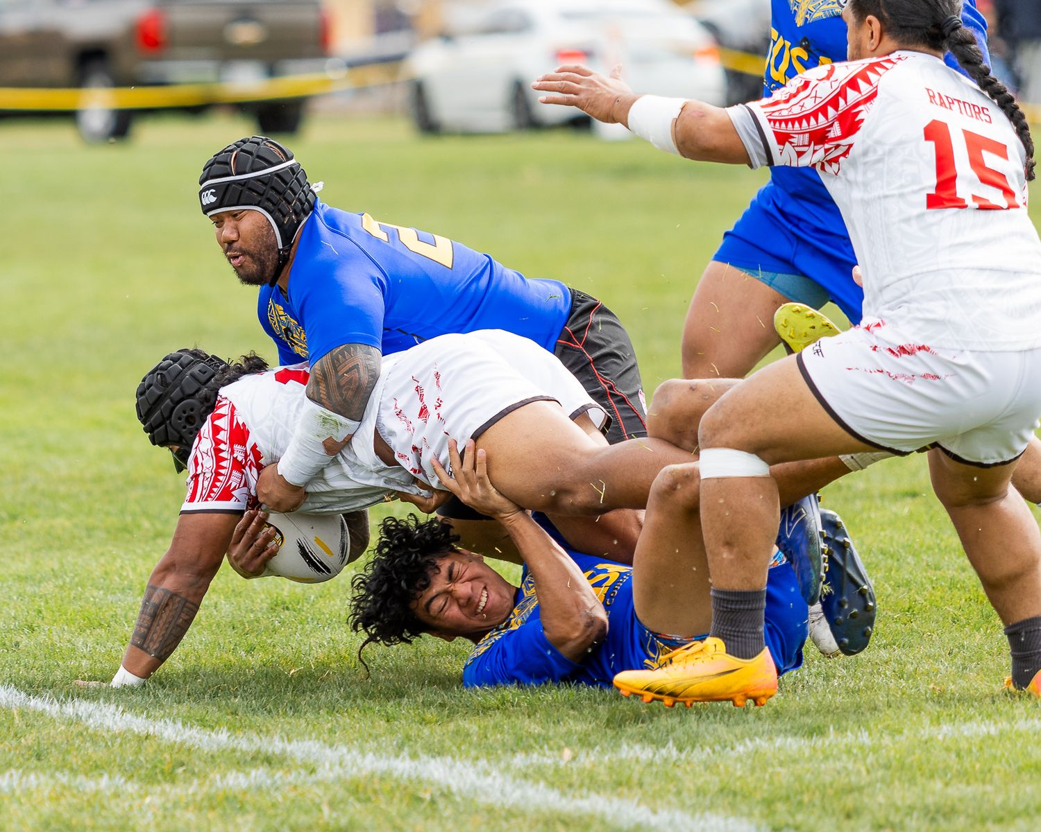 Rugby players in blue and red jerseys compete in an intense tackle during a match on a green grass field.