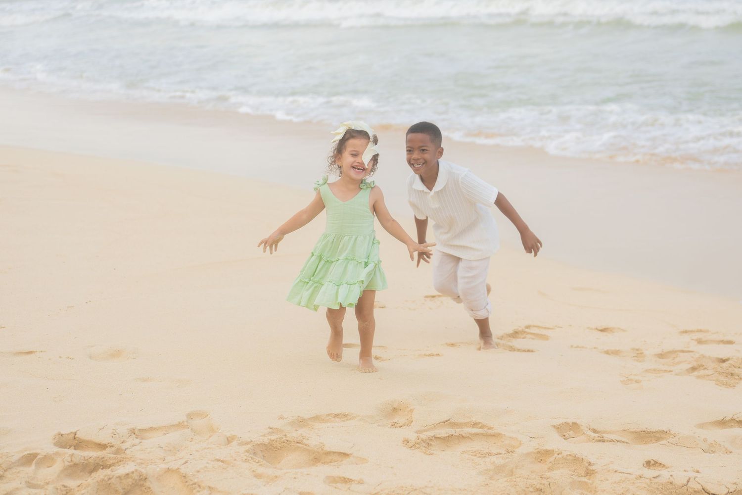 Two happy children running and playing together on a sandy beach with ocean waves in the background.