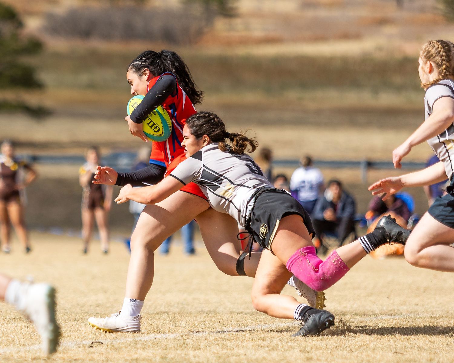 Female rugby player in colorful jersey breaks free from a diving tackle during an outdoor match on dry grass.