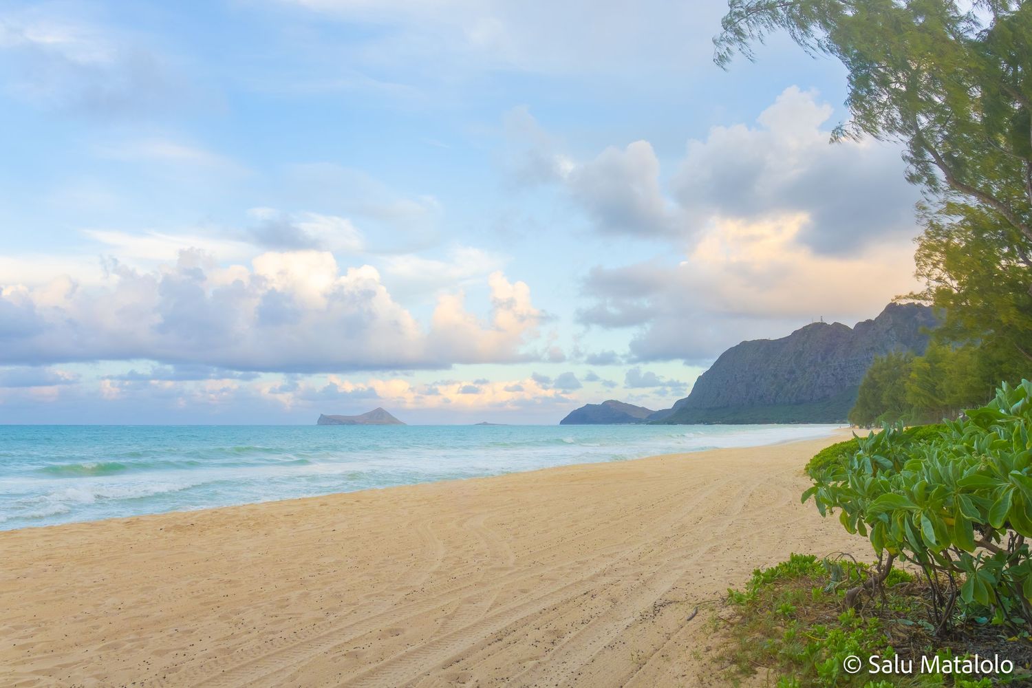 Scenic view of a sandy Hawaiian beach with turquoise ocean waves, coastal greenery, and distant mountains under a cloudy sky.