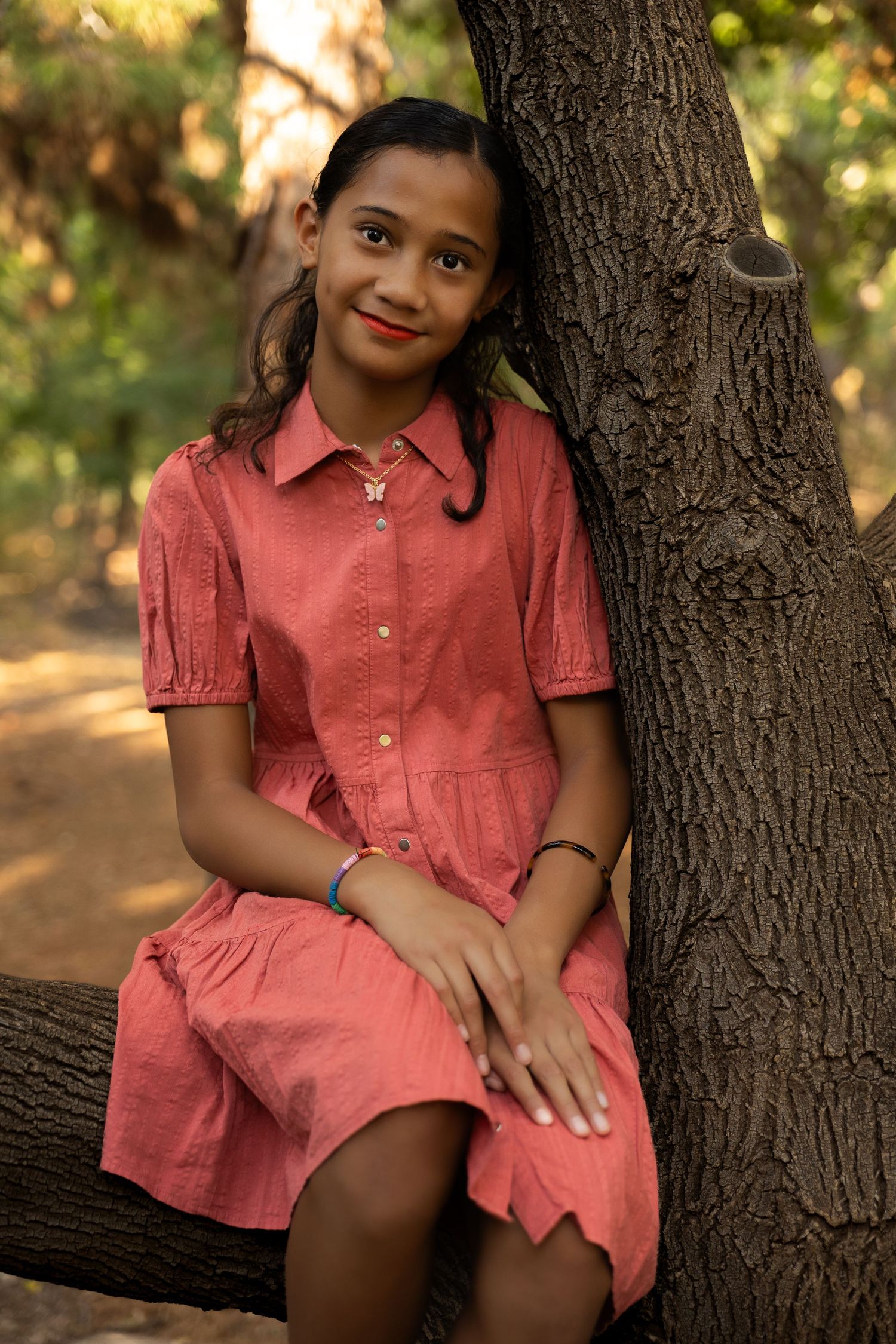 Young girl in pink dress sitting against a tree trunk in a forest, smiling at the camera.