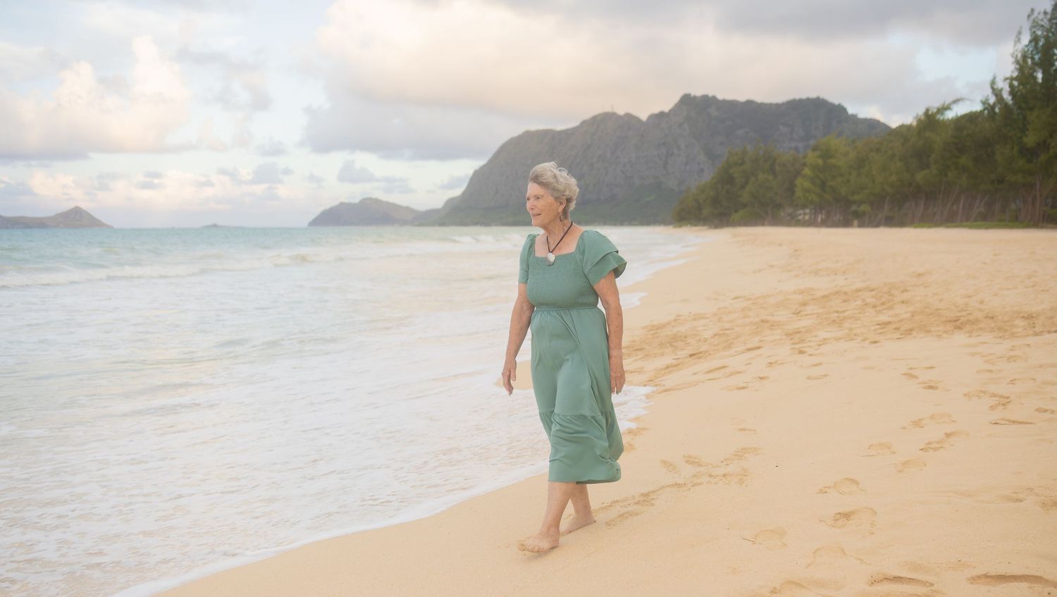 Woman in teal dress walking barefoot on sandy tropical beach with mountains and ocean in background.