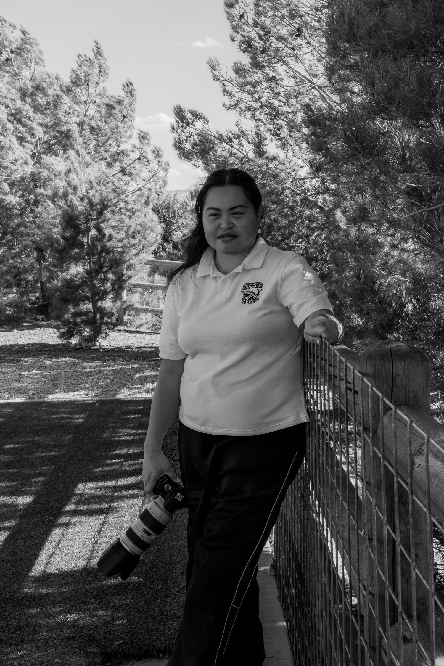 Black and white photo of a woman holding a camera lens, leaning on a fence outdoors surrounded by trees.