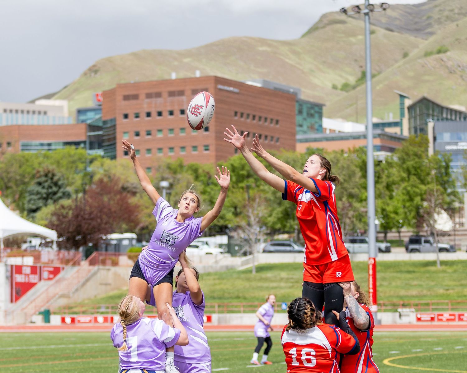 Female rugby players leap for the ball during a lineout in a competitive match with mountains in the background.