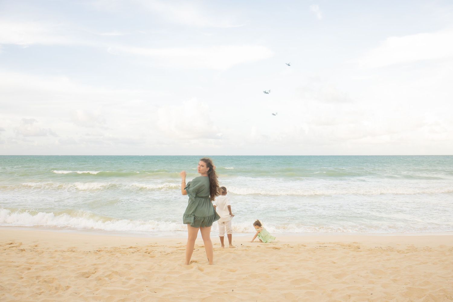 A woman and two children play on a sandy beach with turquoise ocean waves and a cloudy sky backdrop.