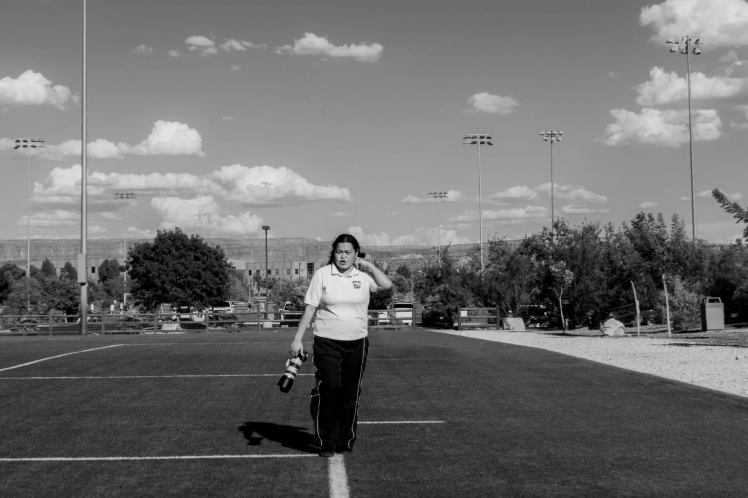 Woman walking on sports field with stadium lights and cloudy sky in black and white photo.
