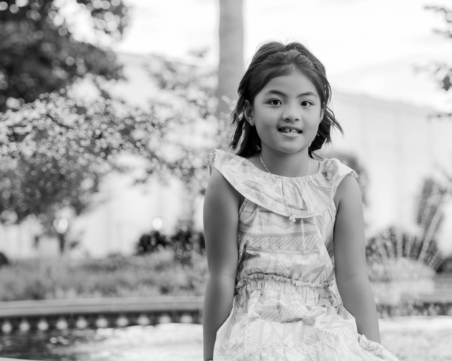 Black and white portrait of a smiling young girl in a ruffled dress with a fountain in the background.