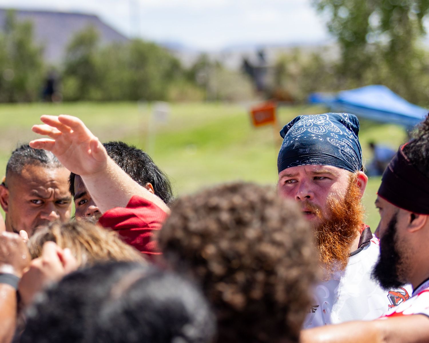 Group of men outdoors at a sports event, featuring a bearded man in a blue bandana and white jersey.