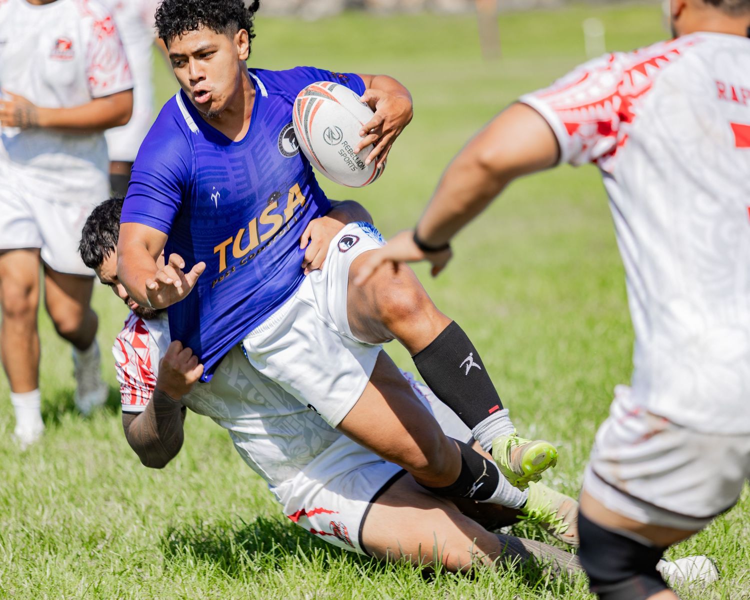 Rugby player in blue Tusa jersey carries ball while being tackled during an outdoor match.