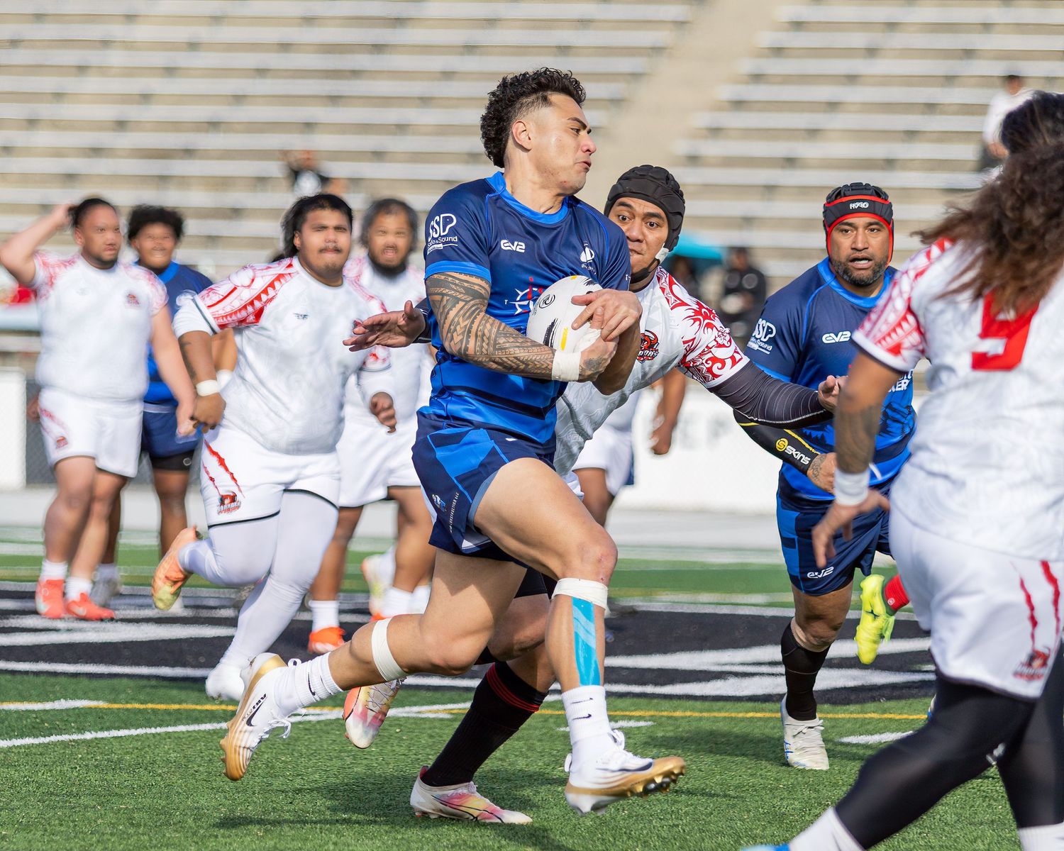 Rugby player in blue uniform carries the ball through defenders in white during a competitive match on turf.