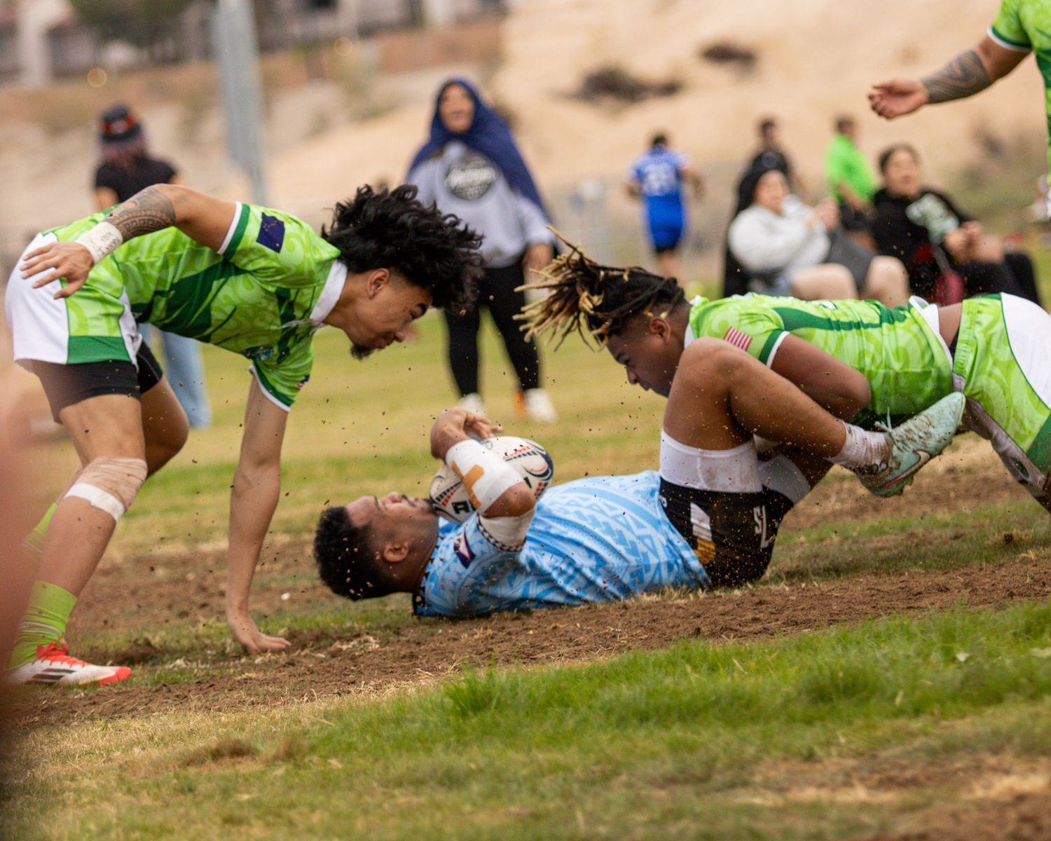 Rugby players in green jerseys tackle an opponent in a light blue jersey during an intense match on a muddy field.