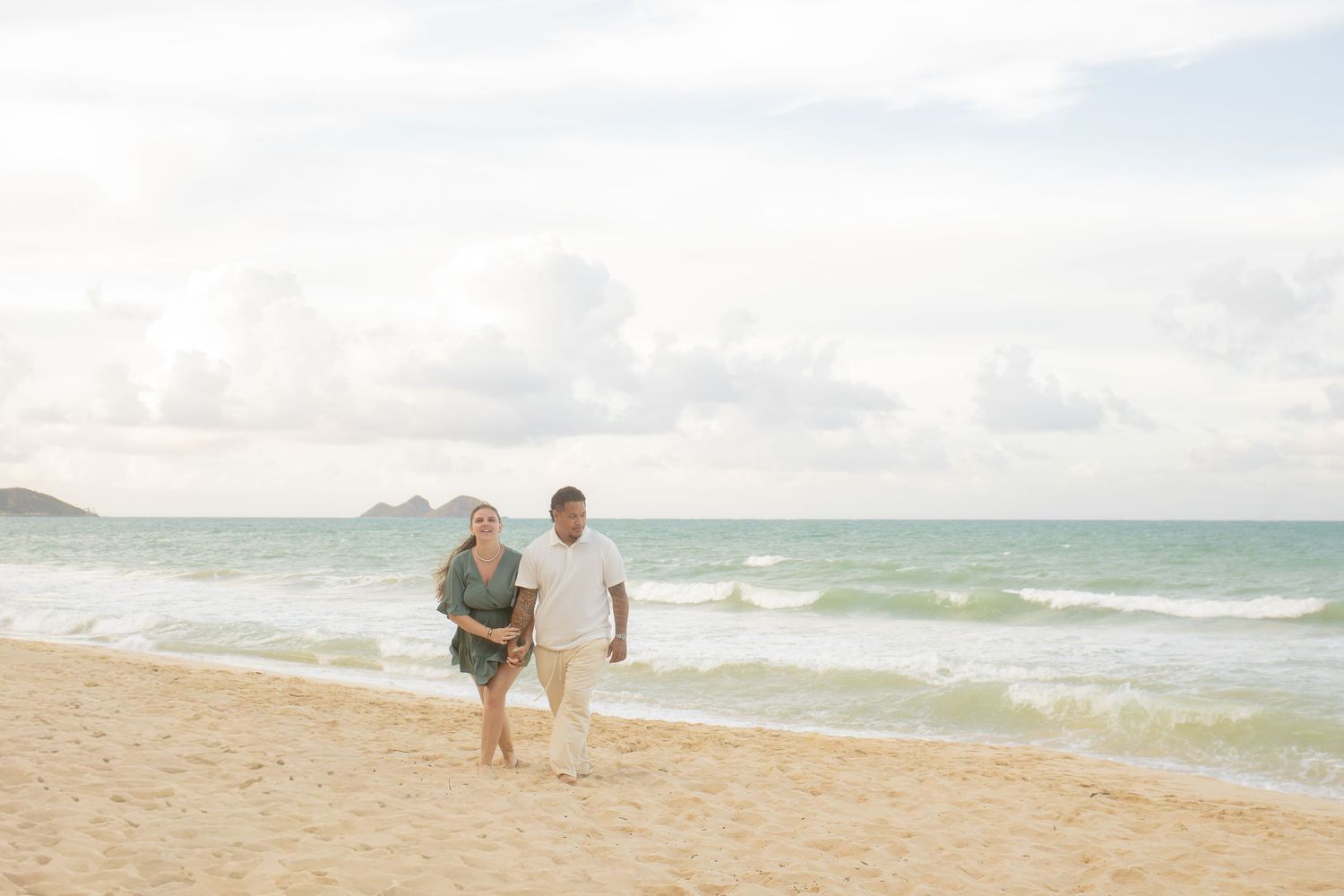 Couple walking hand in hand on a sandy beach with turquoise ocean waves and cloudy sky backdrop.