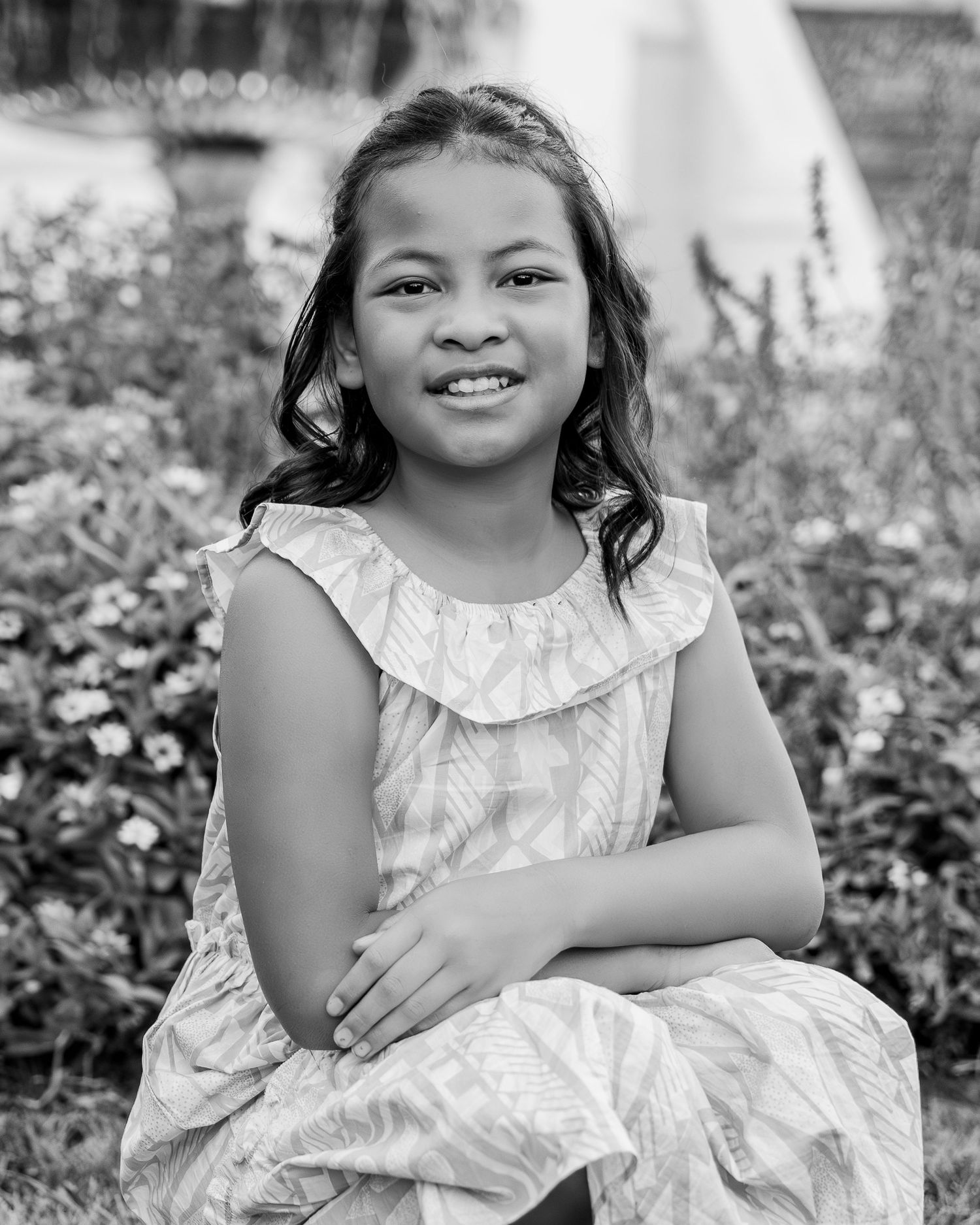 Black and white portrait of a smiling young girl in a ruffled dress, sitting outdoors with flowers in the background.
