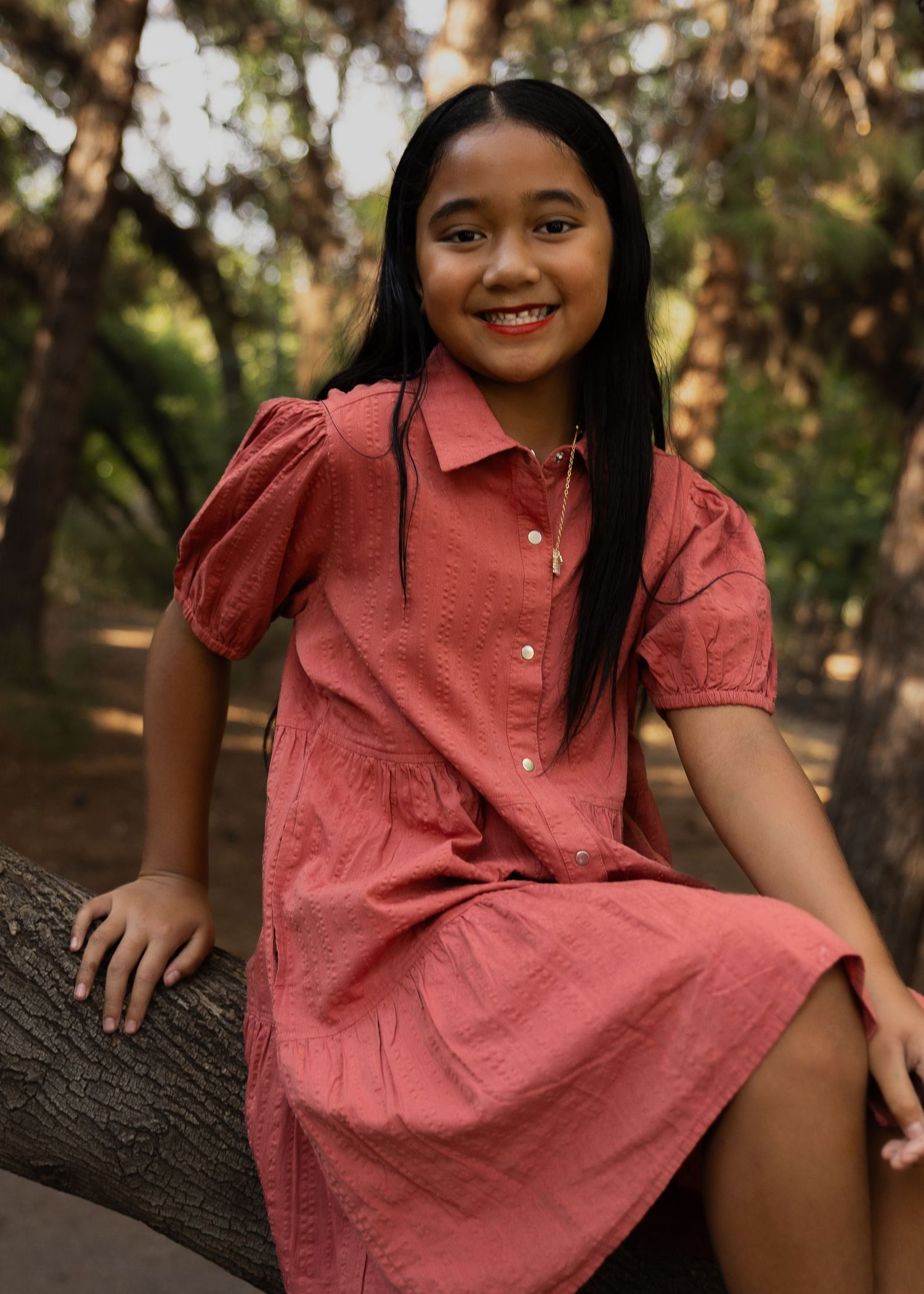 Young girl in a red dress smiling while sitting on a tree branch in a wooded outdoor setting.