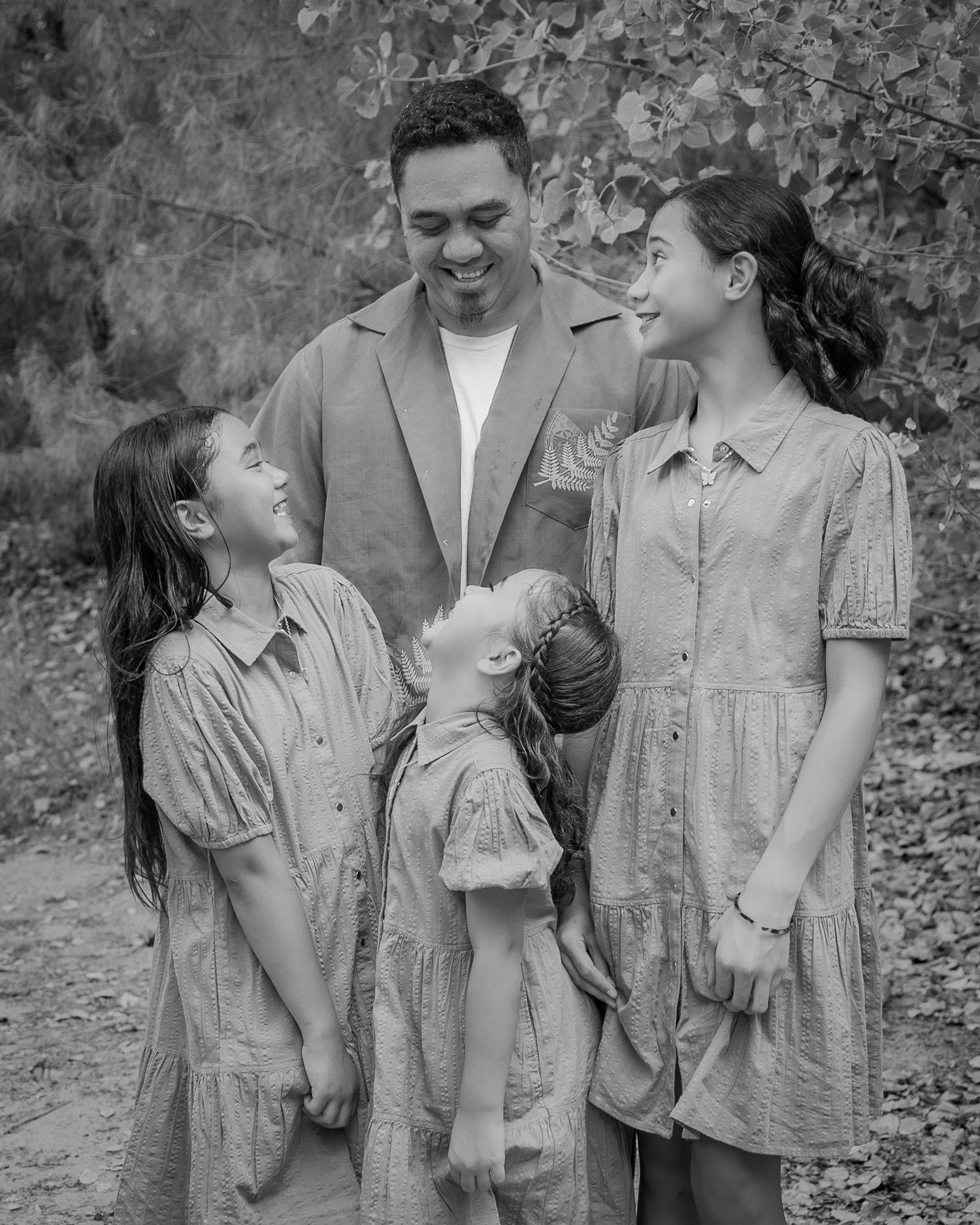 Black and white family portrait of a father smiling with three daughters outdoors among autumn foliage.