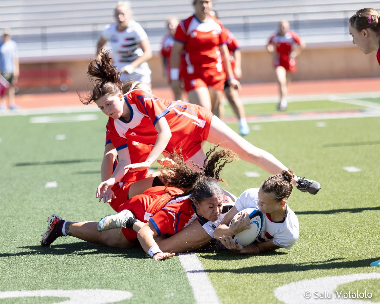 Women's rugby players in red and white uniforms compete in an intense tackle during an outdoor match on a green field.
