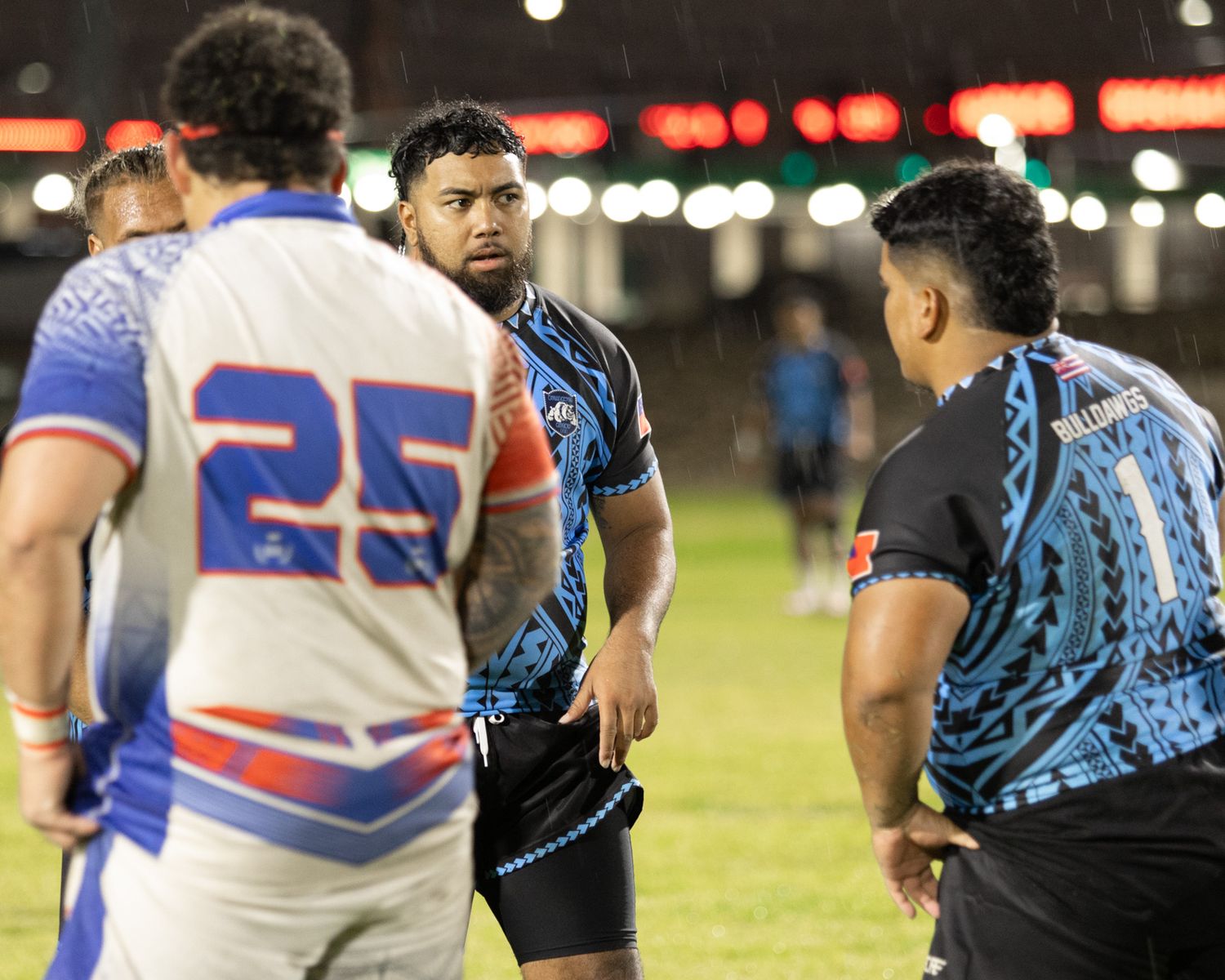 Rugby league players in blue and white jerseys huddle during a night match on a wet field.