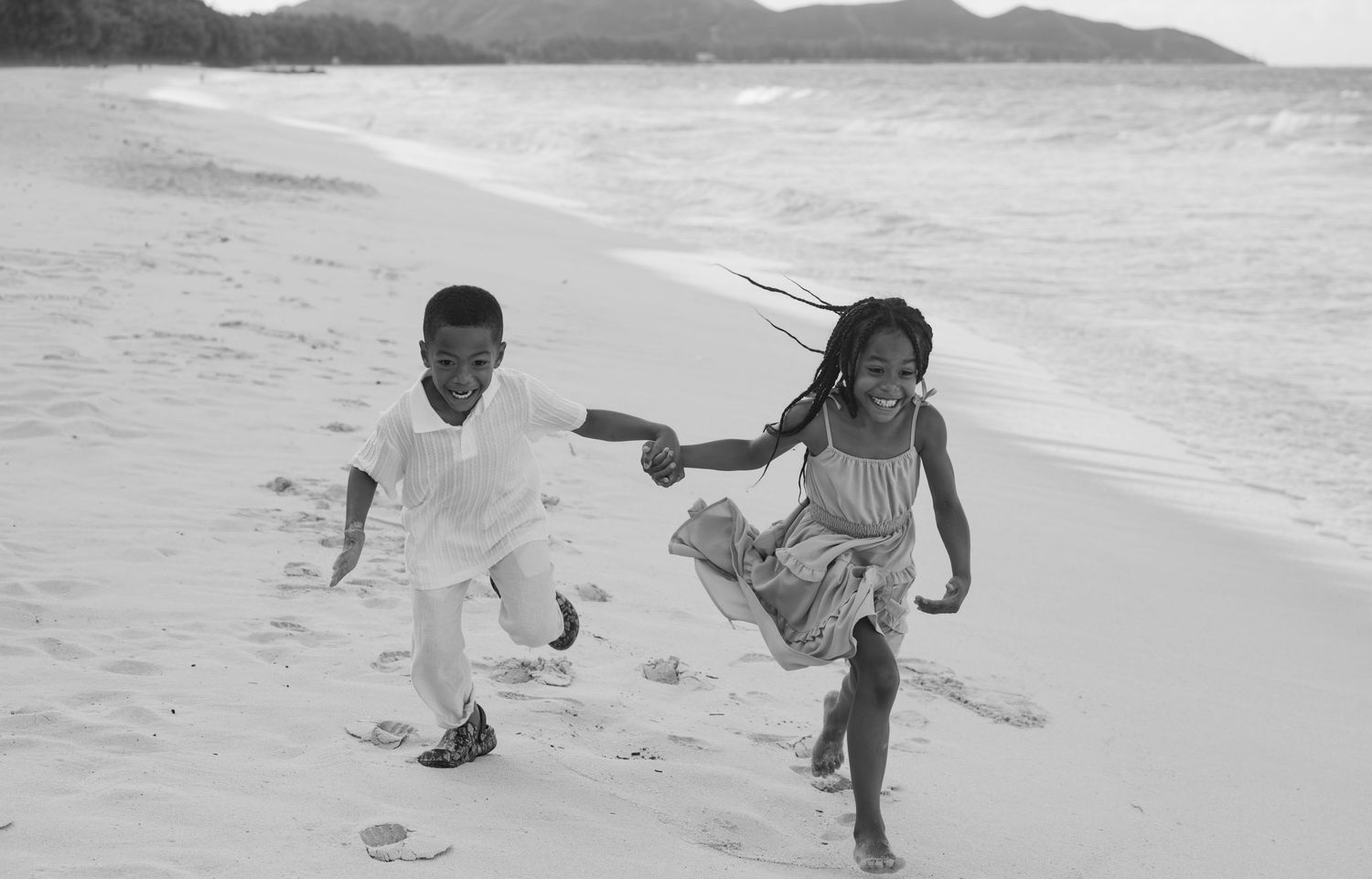 Two children laughing and running together on a sandy beach in a black and white photo.