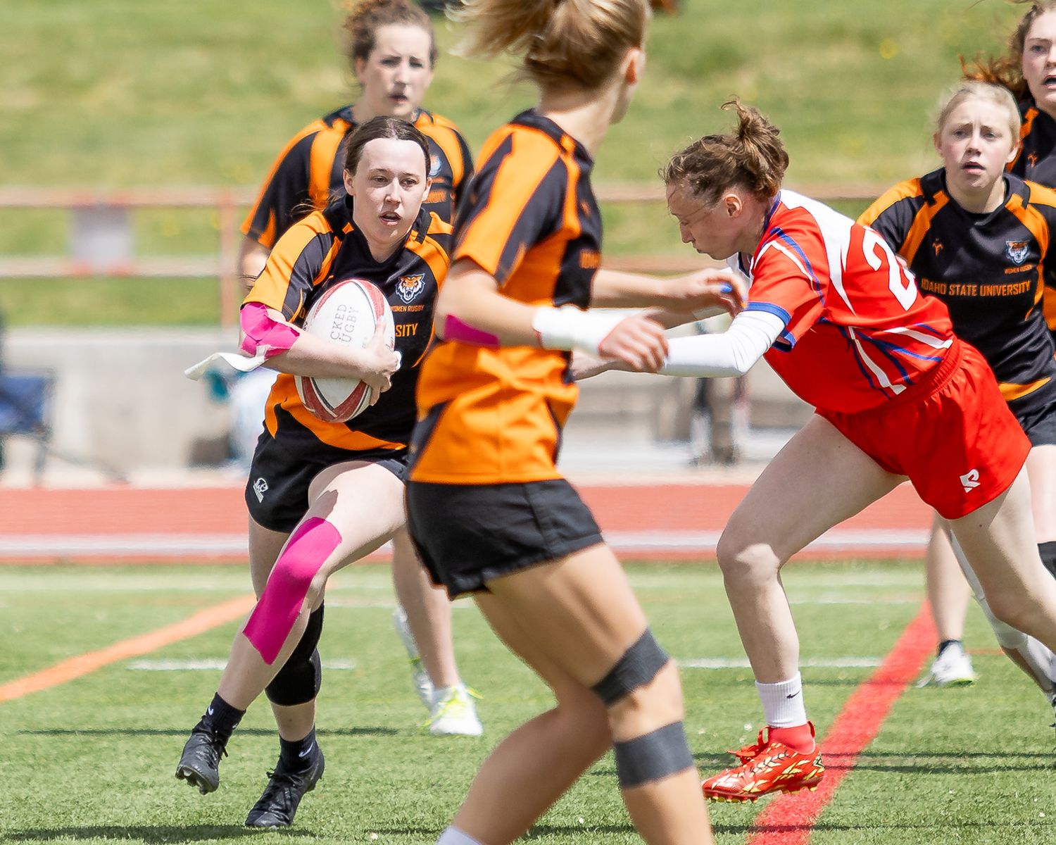 Women's rugby players in black and orange uniforms compete against red team during an outdoor match on turf field.