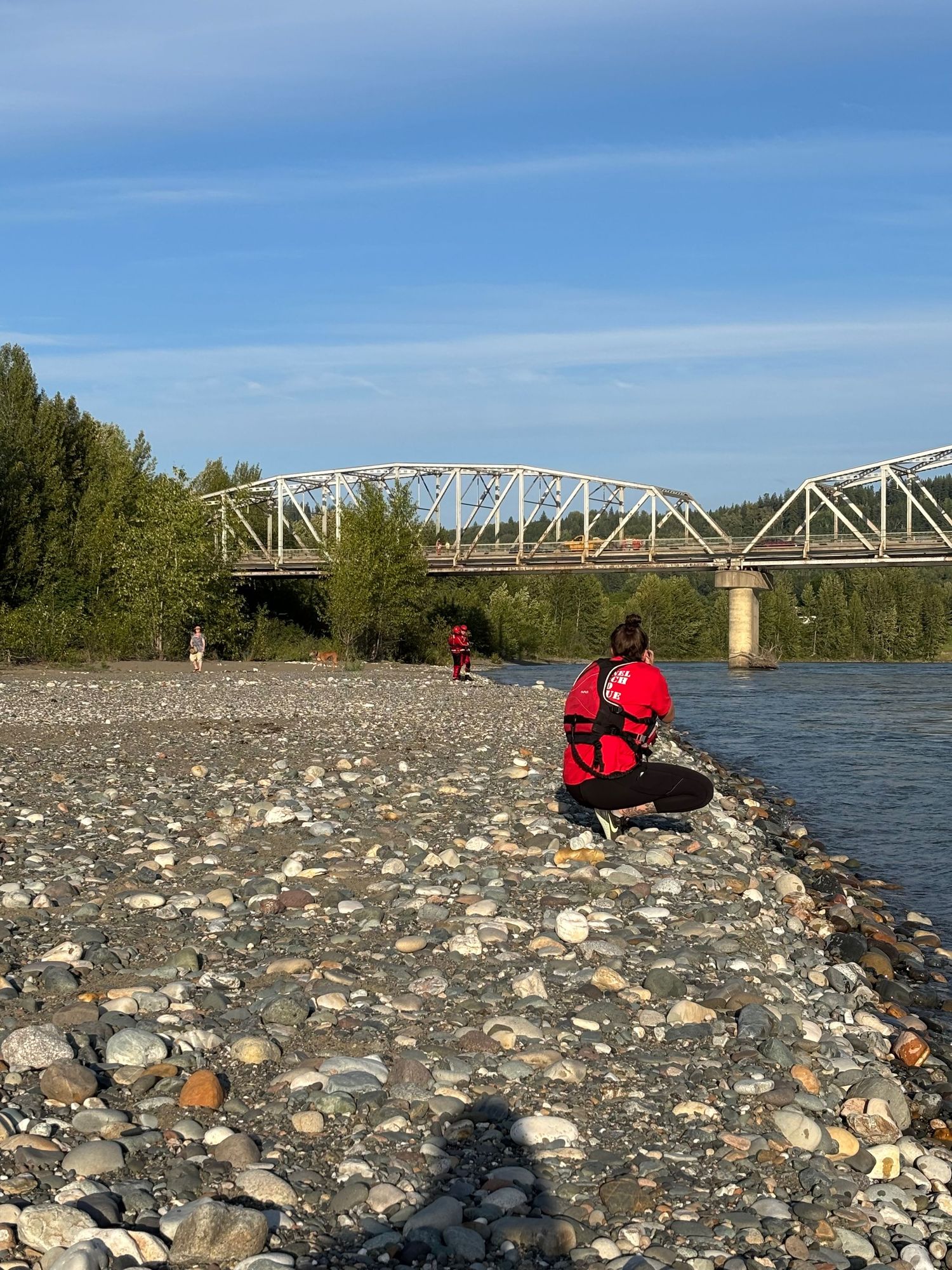A person in red sits on a rocky riverbank overlooking a steel bridge spanning across the water on a sunny day.