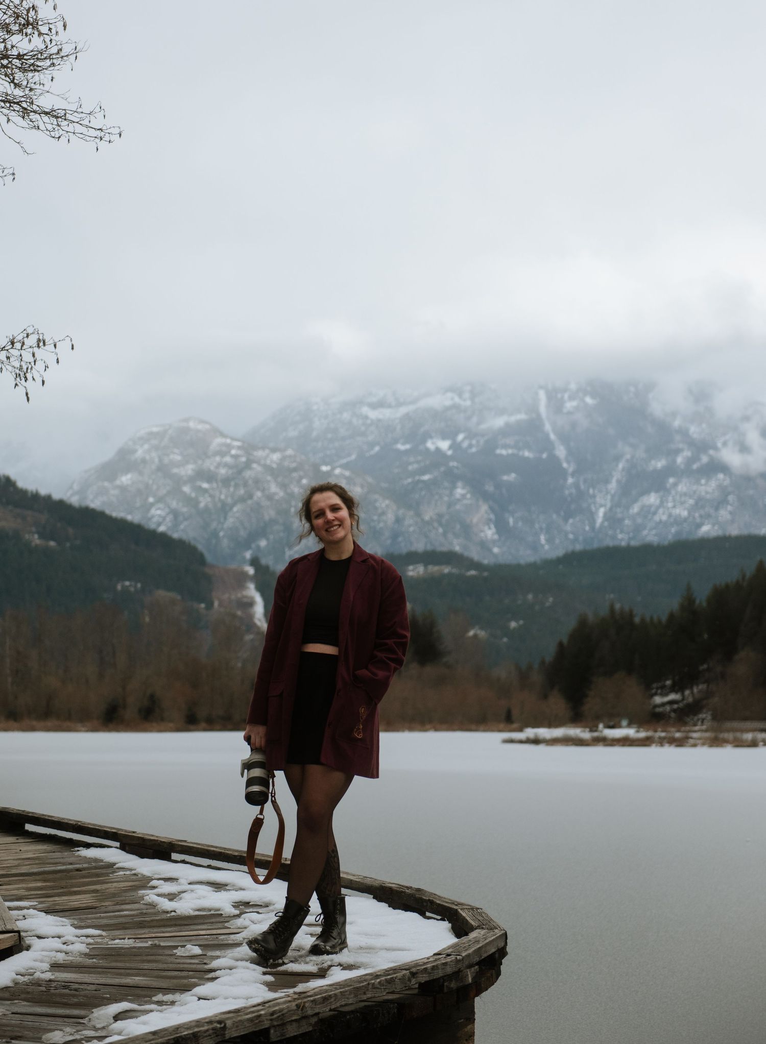 Woman in red coat stands on snowy dock with frozen lake and snow-capped mountains in background.