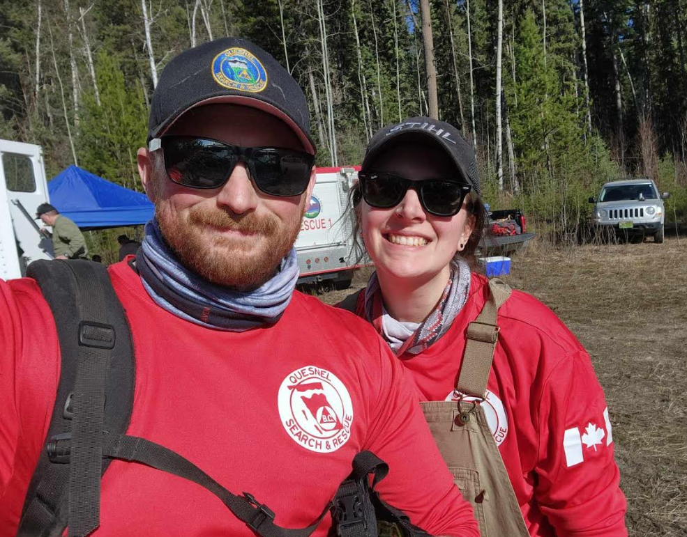 Two outdoor volunteers in red shirts and baseball caps smile together at a wilderness campsite with trees in background.