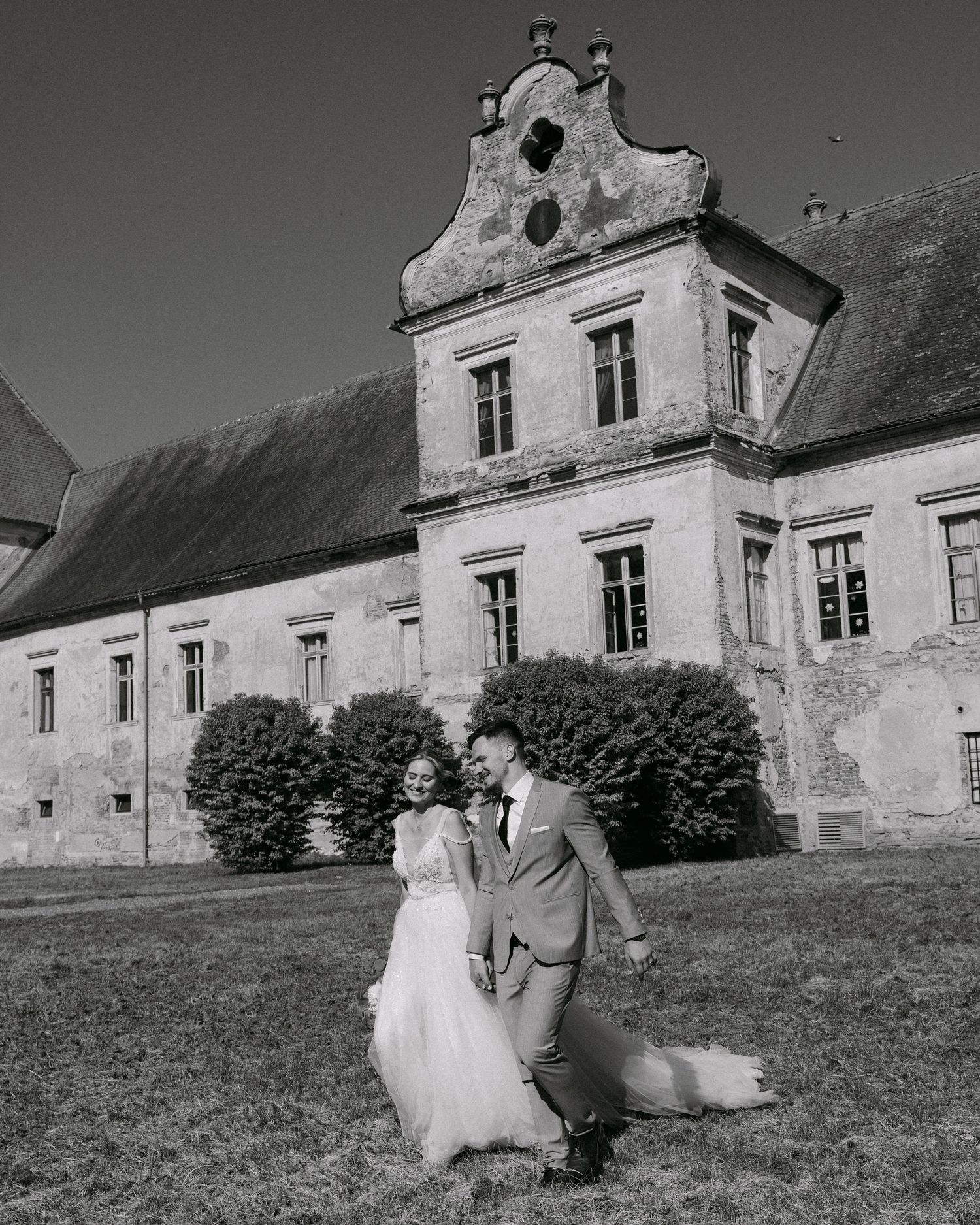 A dramatic monochrome portrait of a newly married couple walking along the sprawling lawns of historic Murska Sobota Castle.